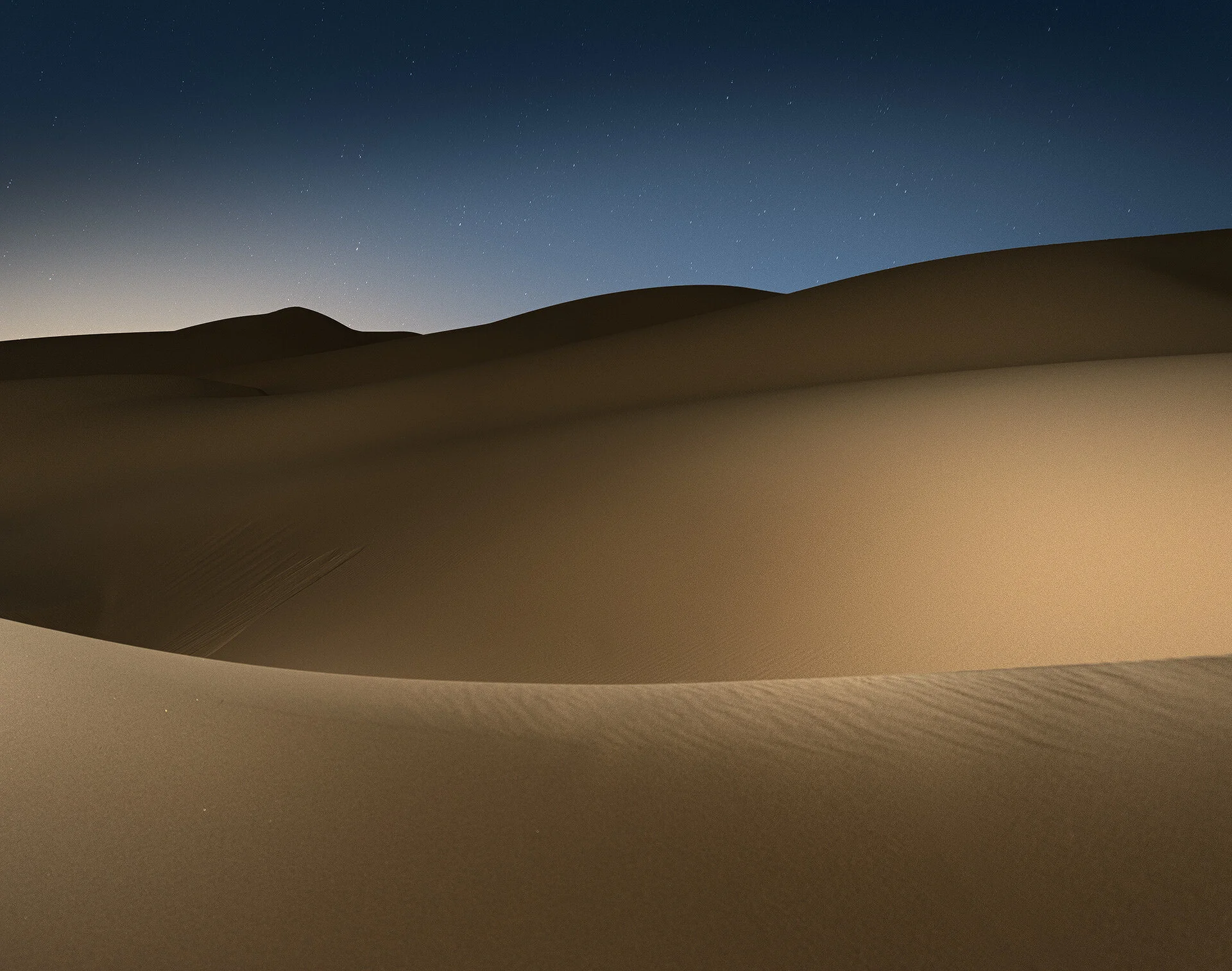 Night photo of smooth rolling sand dune crests illuminated by remote flash against dark blue sky by Hammad Iqbal