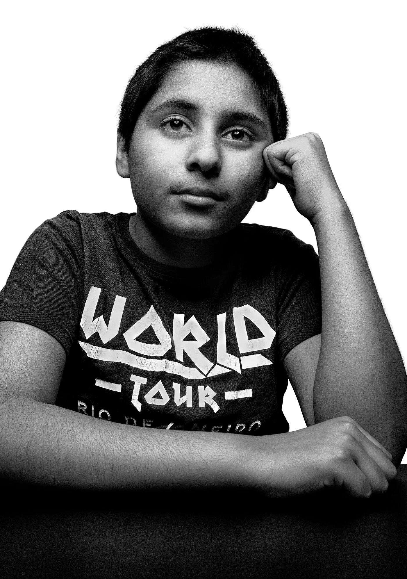 Black and white portrait of young boy leaning at table in World Tour t-shirt by Hammad Iqbal