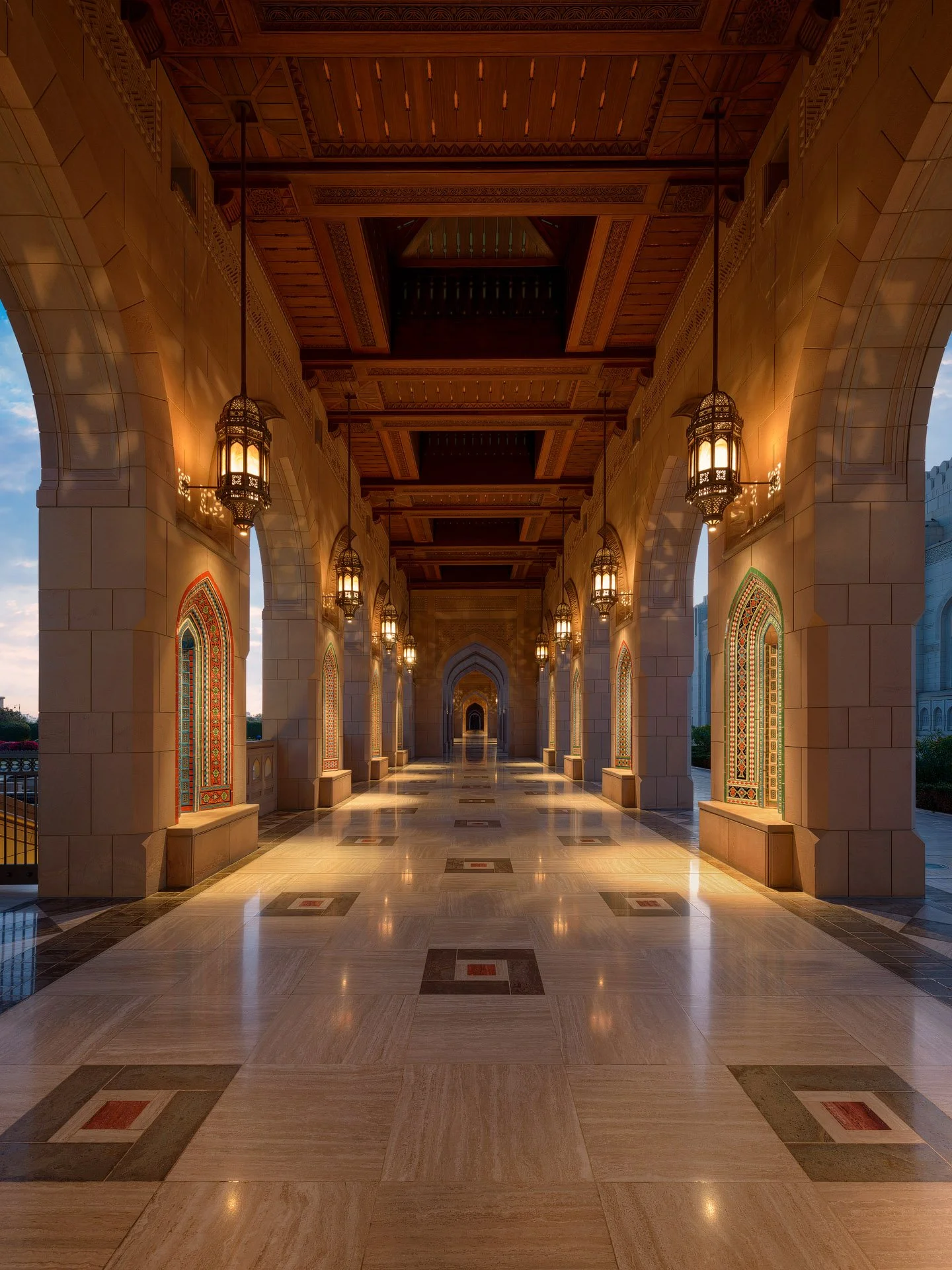 Symmetrical twilight photo looking down an illuminated outdoor arcade corridor with carved wooden ceilings at Sultan Qaboos Grand Mosque in Muscat Oman by Hammad Iqbal