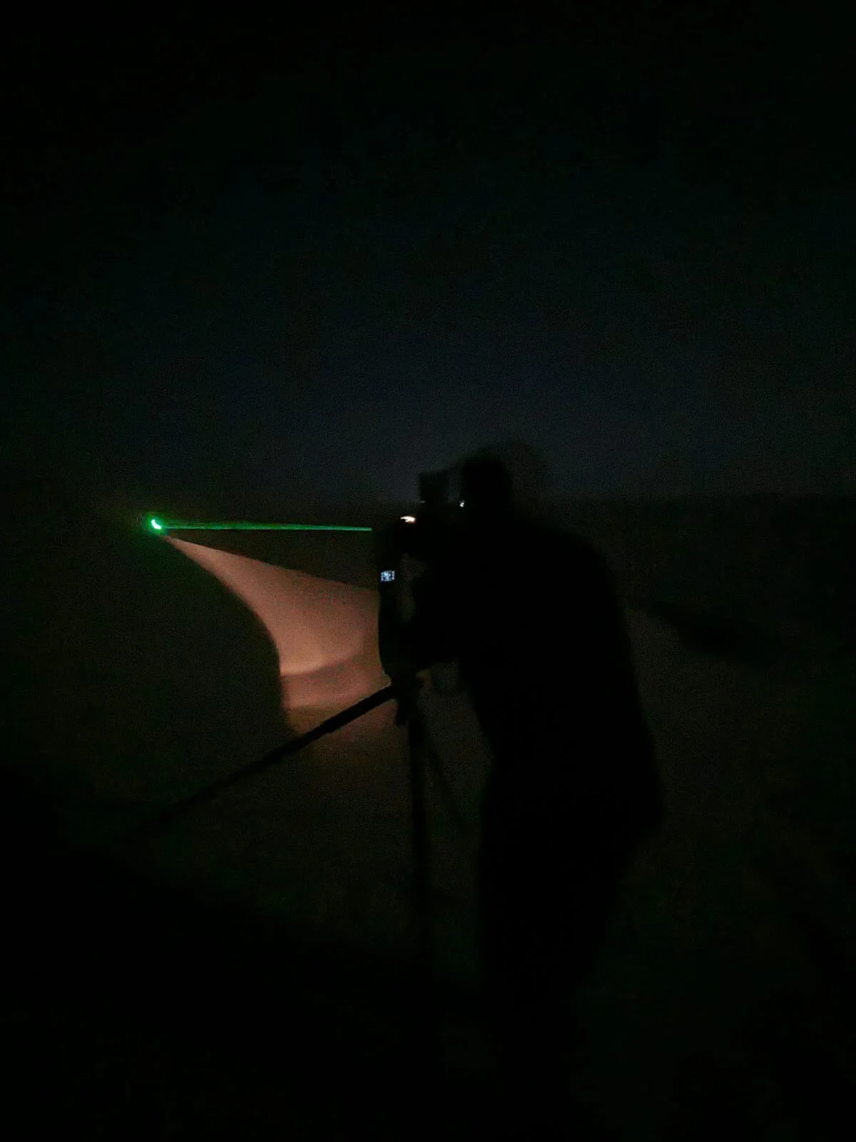 Behind the scenes view of green laser beam illuminating target sand dune ridge for manual camera focusing at night