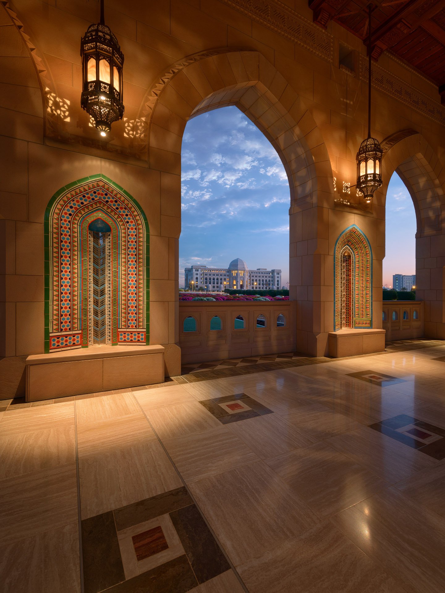 Vertical twilight photograph looking through a sandstone archway with decorative mosaic niches and lanterns at Sultan Qaboos Grand Mosque in Muscat Oman by Hammad Iqbal