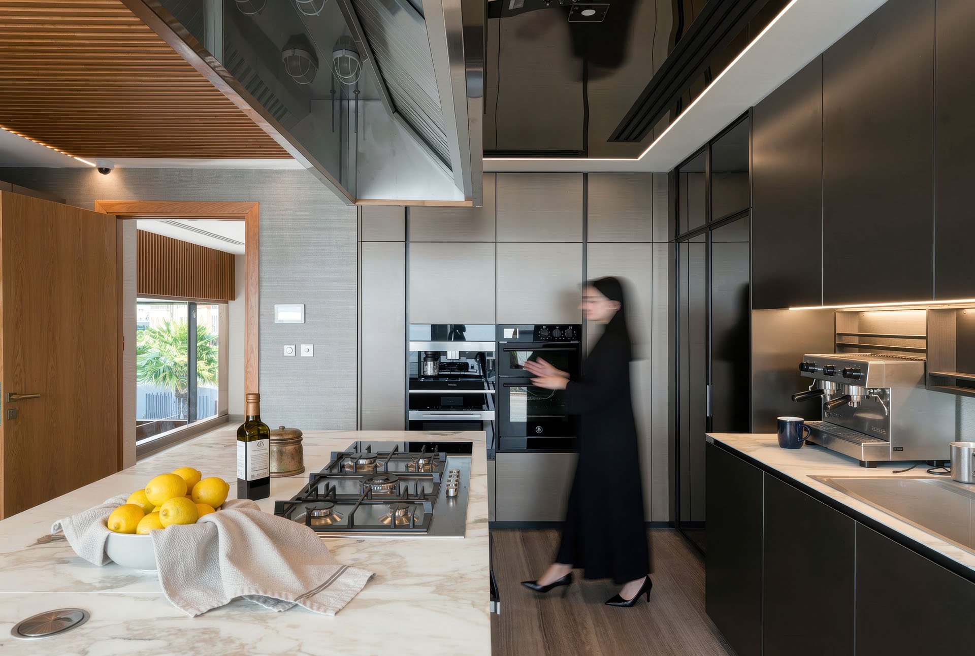 Wide architectural view of a modern dark executive kitchen with marble waterfall island, built-in appliances, and espresso machine in a luxury villa at The Pearl Qatar by Hammad Iqbal