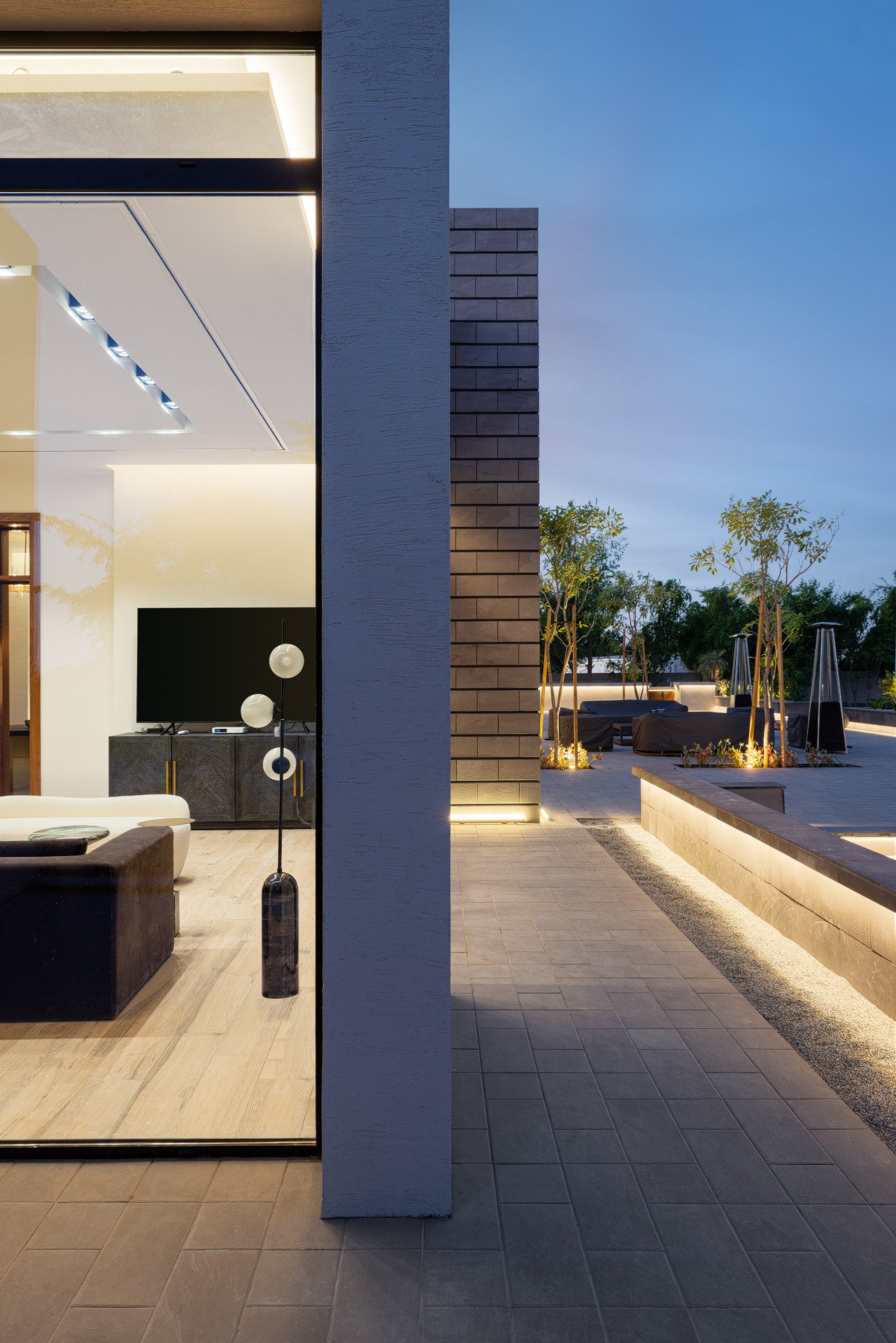 Architectural vignette looking through open glass sliding door past textured stone pillar onto illuminated terrace by Hammad Iqbal