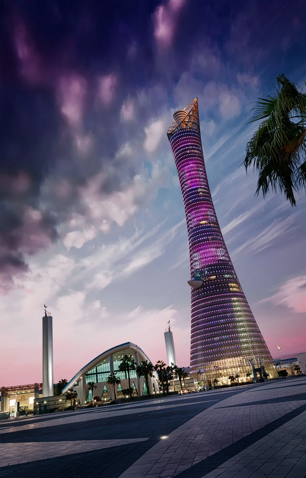 Low-angle evening architectural photography of the illuminated 300-meter Torch Doha hotel glowing in vibrant purple LEDs beside the modern Hilal bin Jaham Al Kuwari Mosque under a dramatic dusk sky.