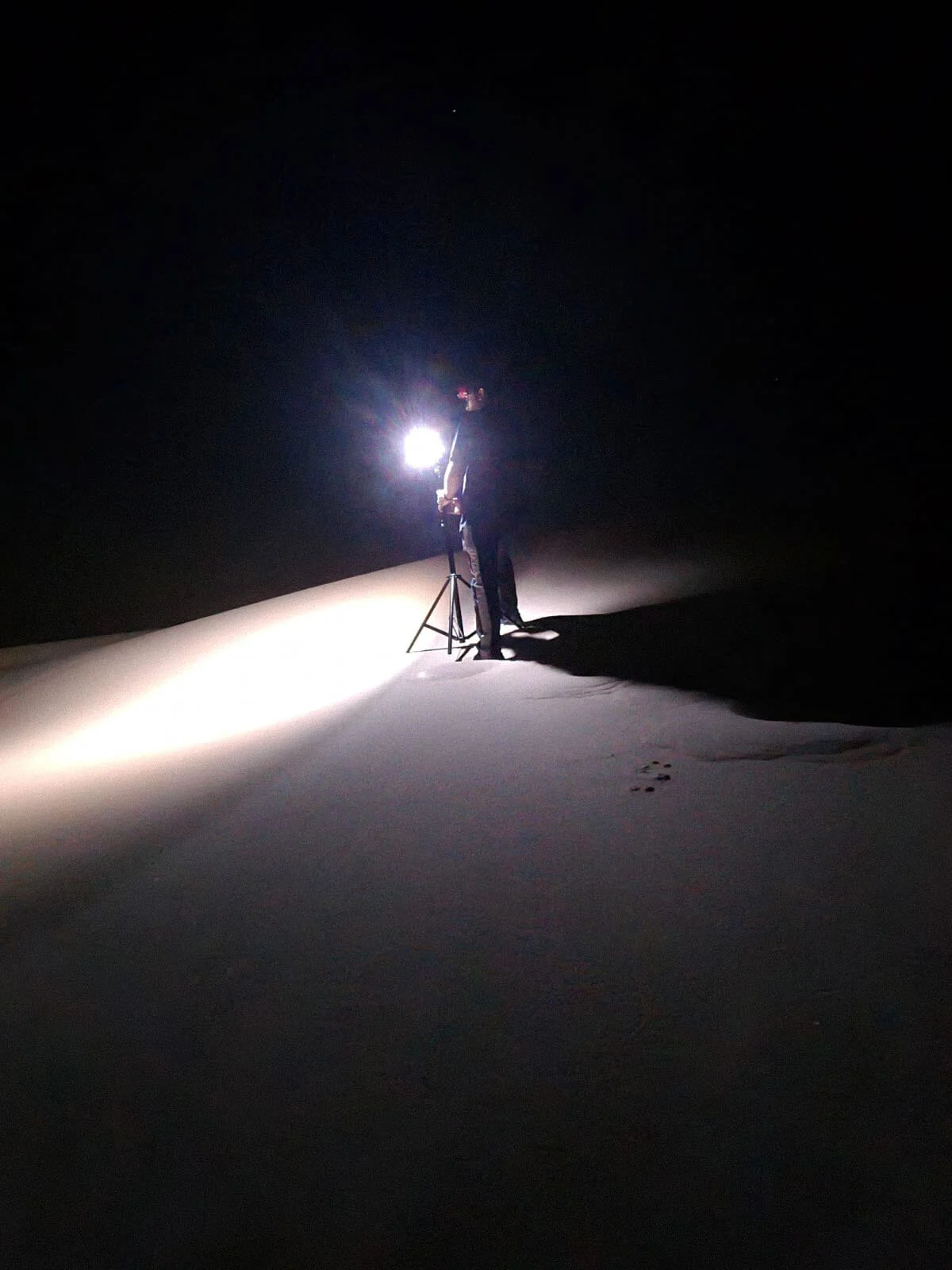 Behind the scenes photograph capturing off camera strobe flash firing to illuminate texture on desert sand dunes at night