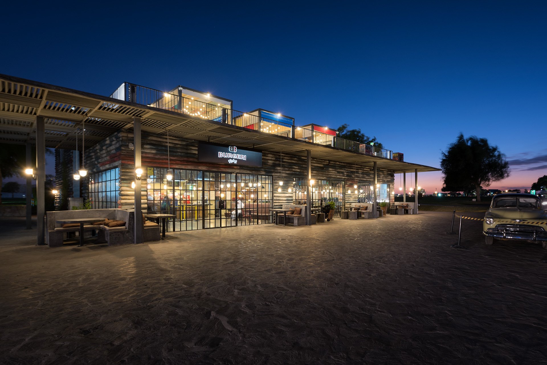 Twilight exterior architectural photograph of Burgeri restaurant in Aspire Park, showcasing industrial vintage-style wooden cladding, glass grid facade, illuminated patio seating, and rooftop seating structure.