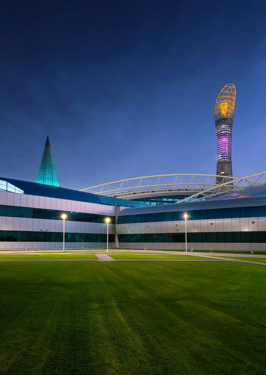 Exterior dusk architectural photograph of the manicured green lawn surrounding Aspire Dome with its illuminated green glass spire, set against the illuminated 300-meter Torch Doha hotel tower under a deep blue evening sky.