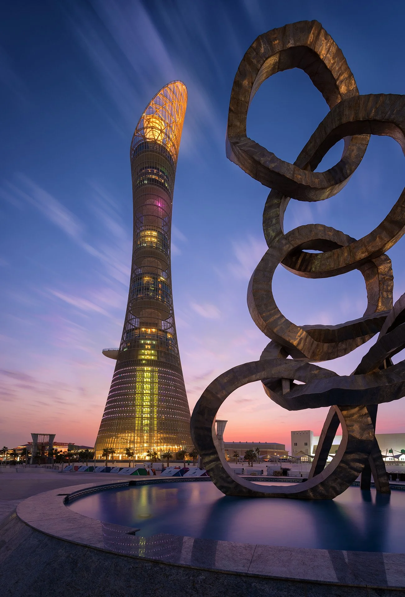Fine art dusk architectural photograph of the illuminated 300-meter Torch Doha hotel tower framed behind the bronze Olympic Rings sculpture and fountain in Aspire Zone, Qatar.