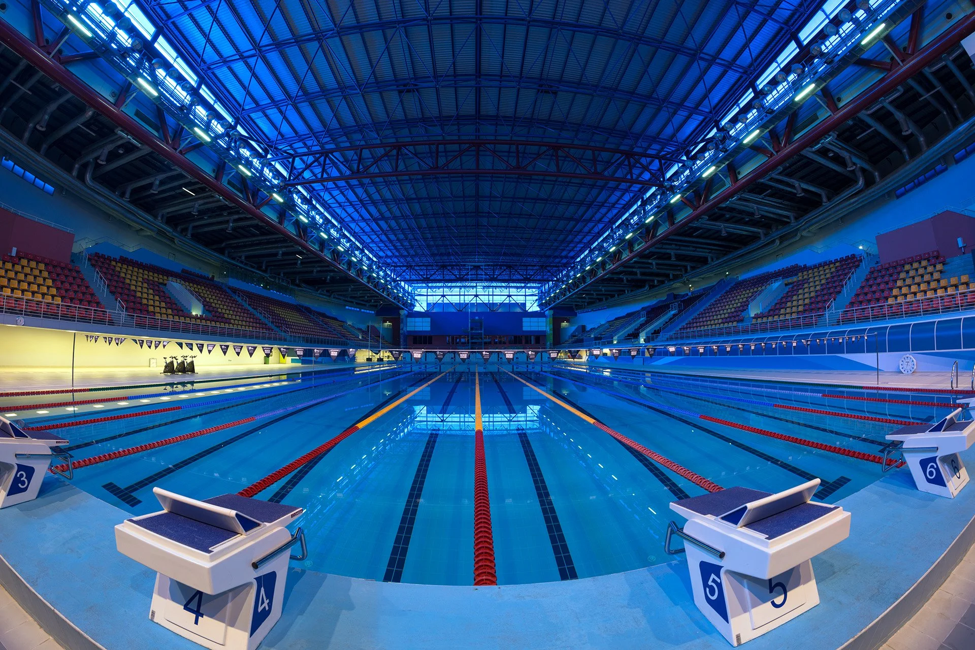 Fisheye interior architectural photography of the Olympic-size swimming pool at Hamad Aquatic Center, showing starting blocks, spectator seating, and blue steel roof structure.