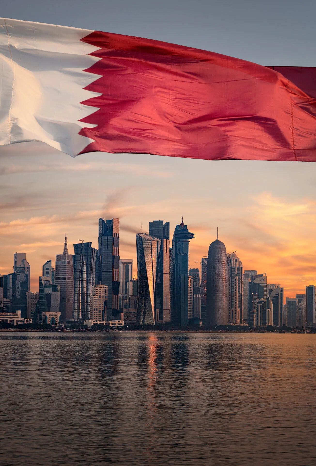 Vertical architectural photo of the Qatari flag unfurled with West Bay high-rise towers in the background by Hammad Iqbal