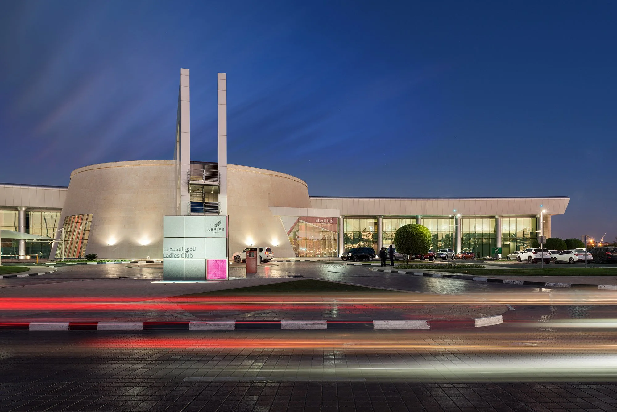 Exterior twilight photograph of the Aspire Ladies Club featuring illuminated stone architecture, glass signage, twin pylons, and long-exposure car light trails in the foreground.