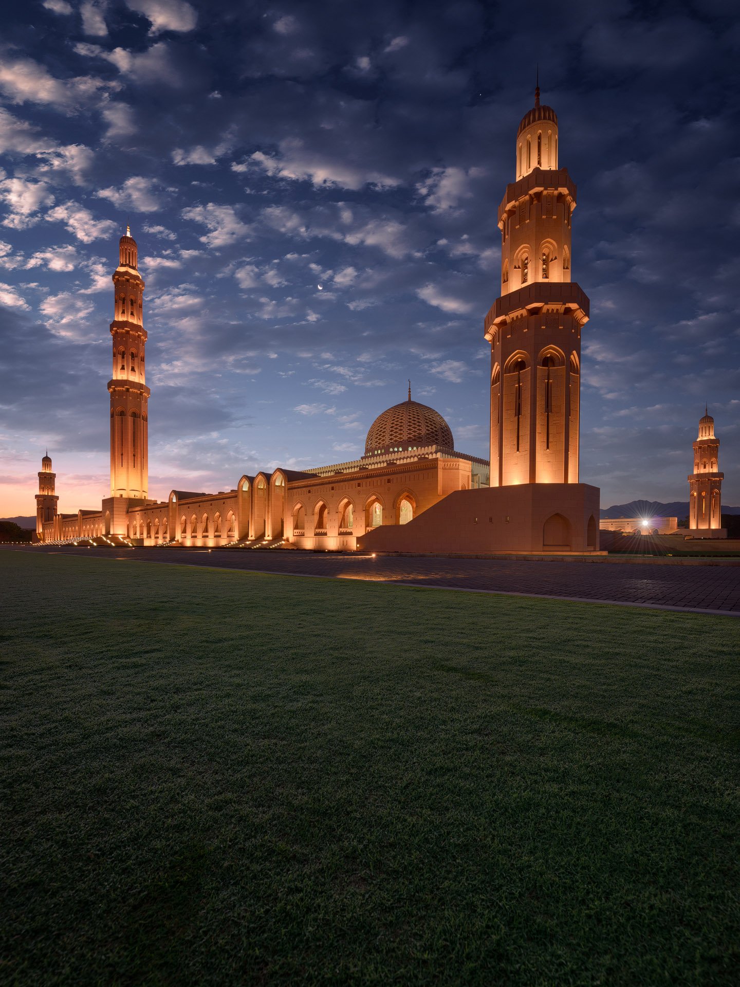 Vertical blue hour exterior photo showing the illuminated sandstone minarets and central dome at Sultan Qaboos Grand Mosque in Muscat Oman by Hammad Iqbal