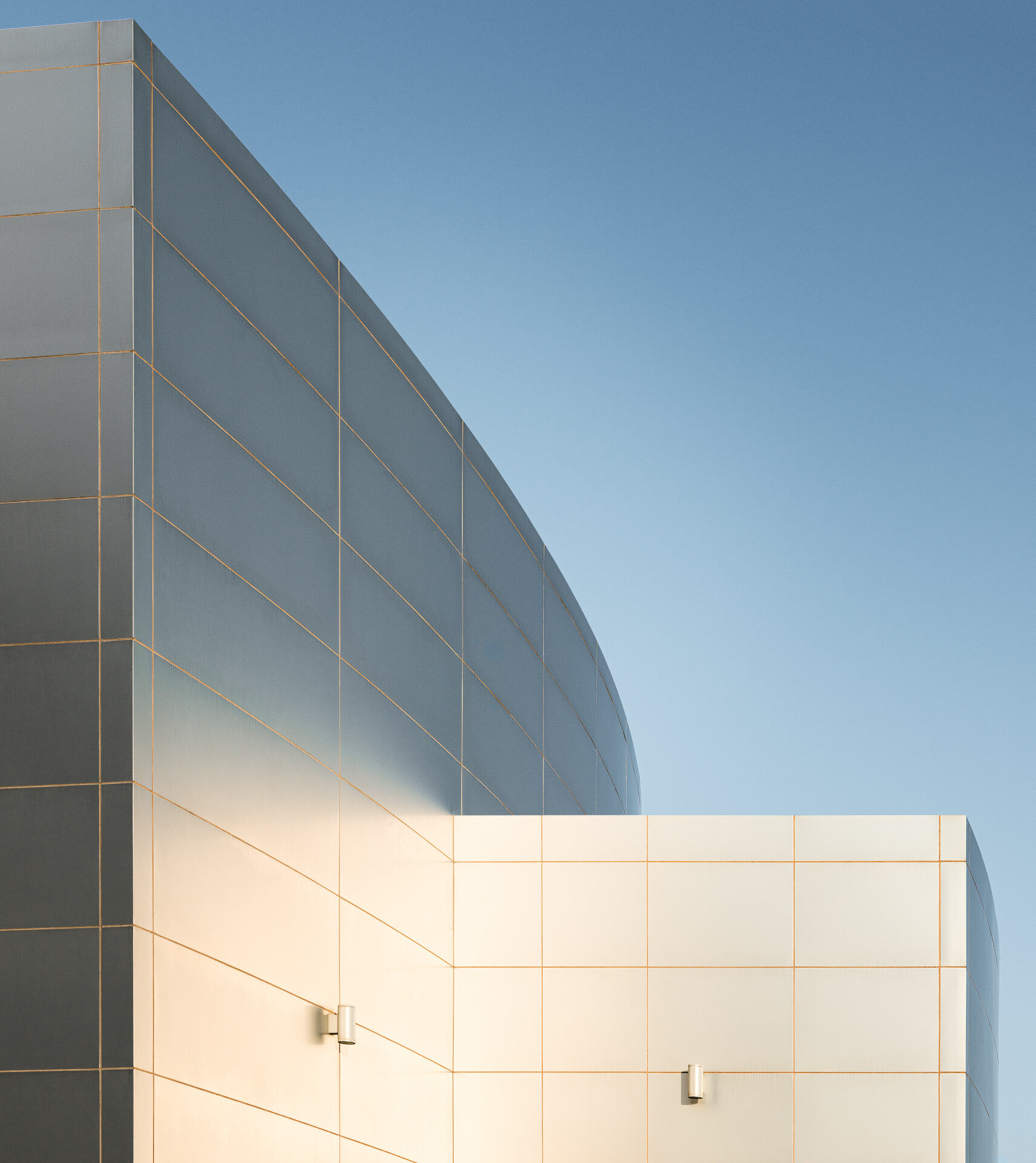 Architectural detail photo showing the curved metallic panel facade and clean geometric lines of the Aspire Zone Ladies Sports Hall against a clear blue sky.
