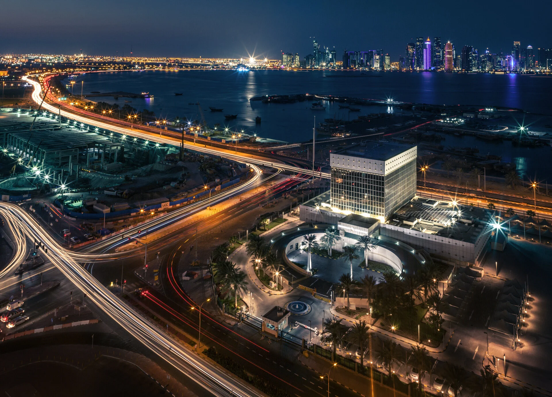 Aerial high angle night photograph of a glowing highway interchange traffic light trails and Doha bay by Hammad Iqbal
