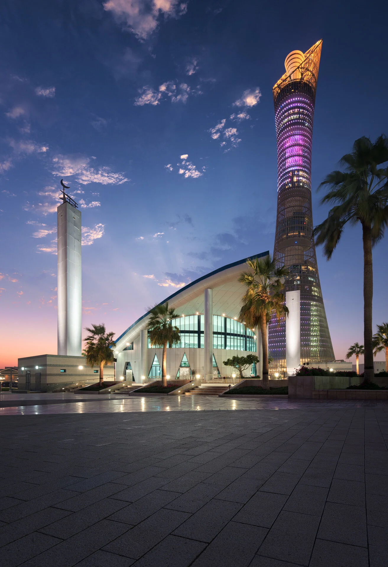 Exterior twilight photography of the Hilal bin Jaham Al Kuwari Mosque with its modern curved roof and standalone minaret, framed alongside the illuminated 300-meter Torch Tower in Aspire Zone, Doha.