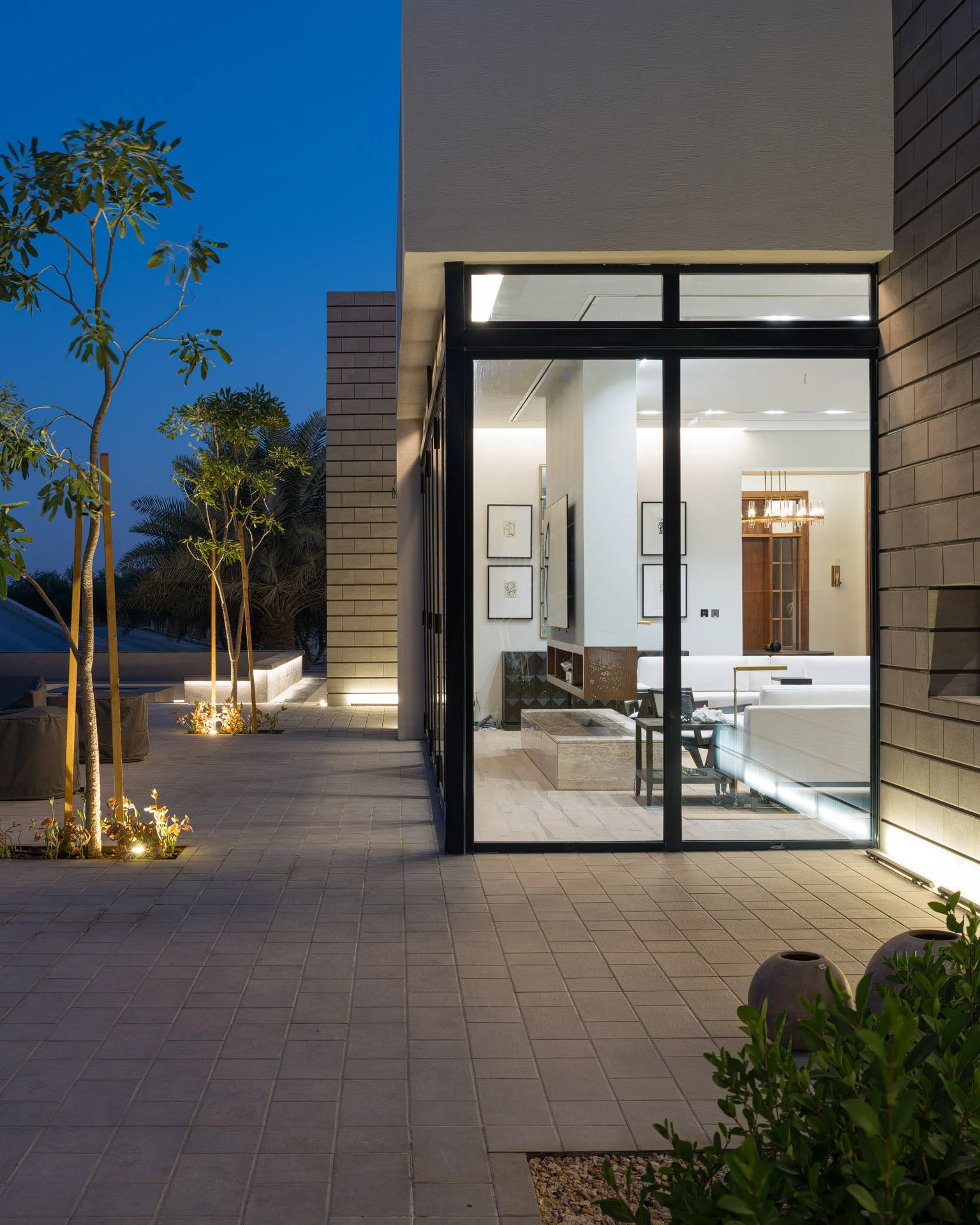 Architectural photo framing illuminated interior living room through floor to ceiling glass corner onto twilight patio in Qatar by Hammad Iqbal