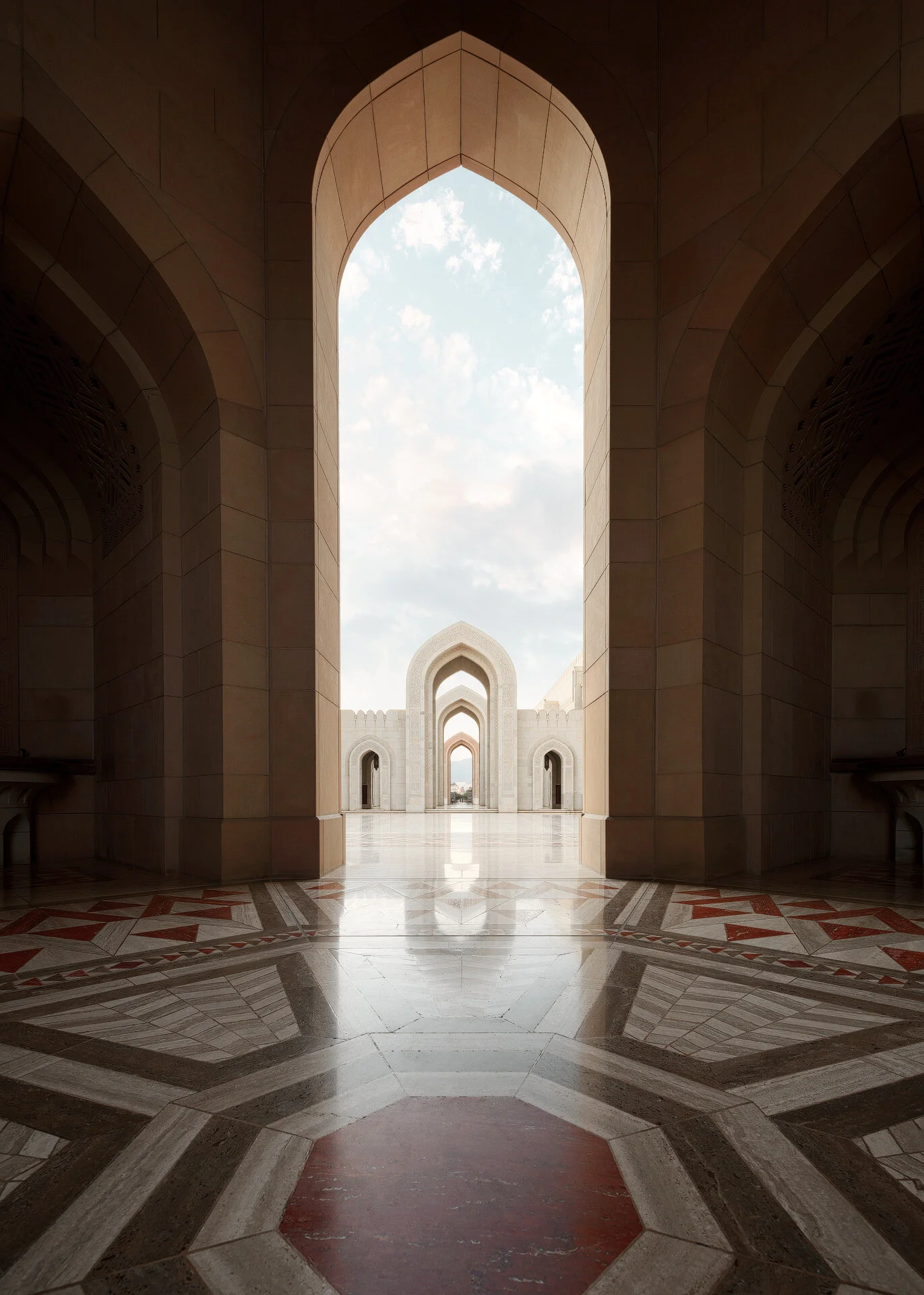 Vertical morning photo looking through sandstone archways across the marble courtyard at Sultan Qaboos Grand Mosque in Muscat Oman by Hammad Iqbal
