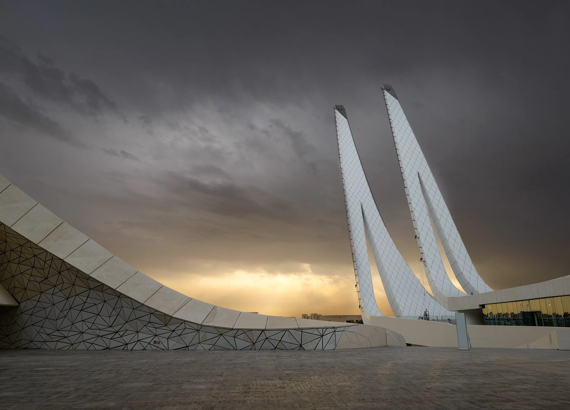 Architectural shot of the Minaretein Center courtyard in Education City under a dramatic storm sky, featuring sweeping curved walls and illuminated twin minarets.