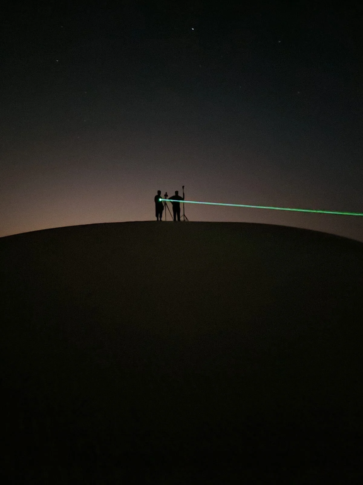 Behind the scenes wide shot showing photographer silhouette on a dune ridge aiming a laser beam for night focus