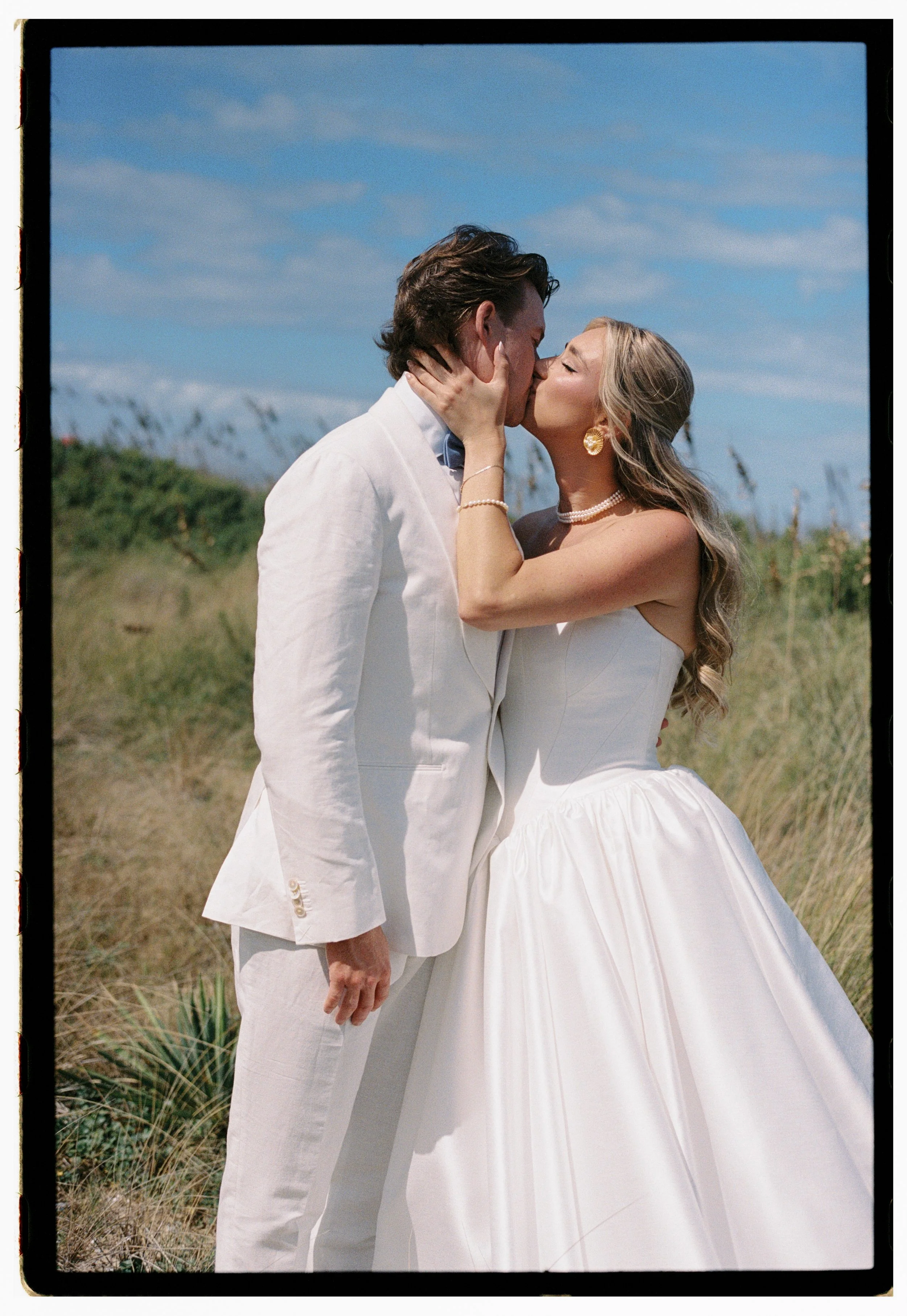 A bride and groom kiss outdoors on a sunny day, with grassy dunes and a blue sky in the background. The bride is wearing a strapless white wedding gown, pearls, and gold earrings, while the groom is dressed in a white suit.