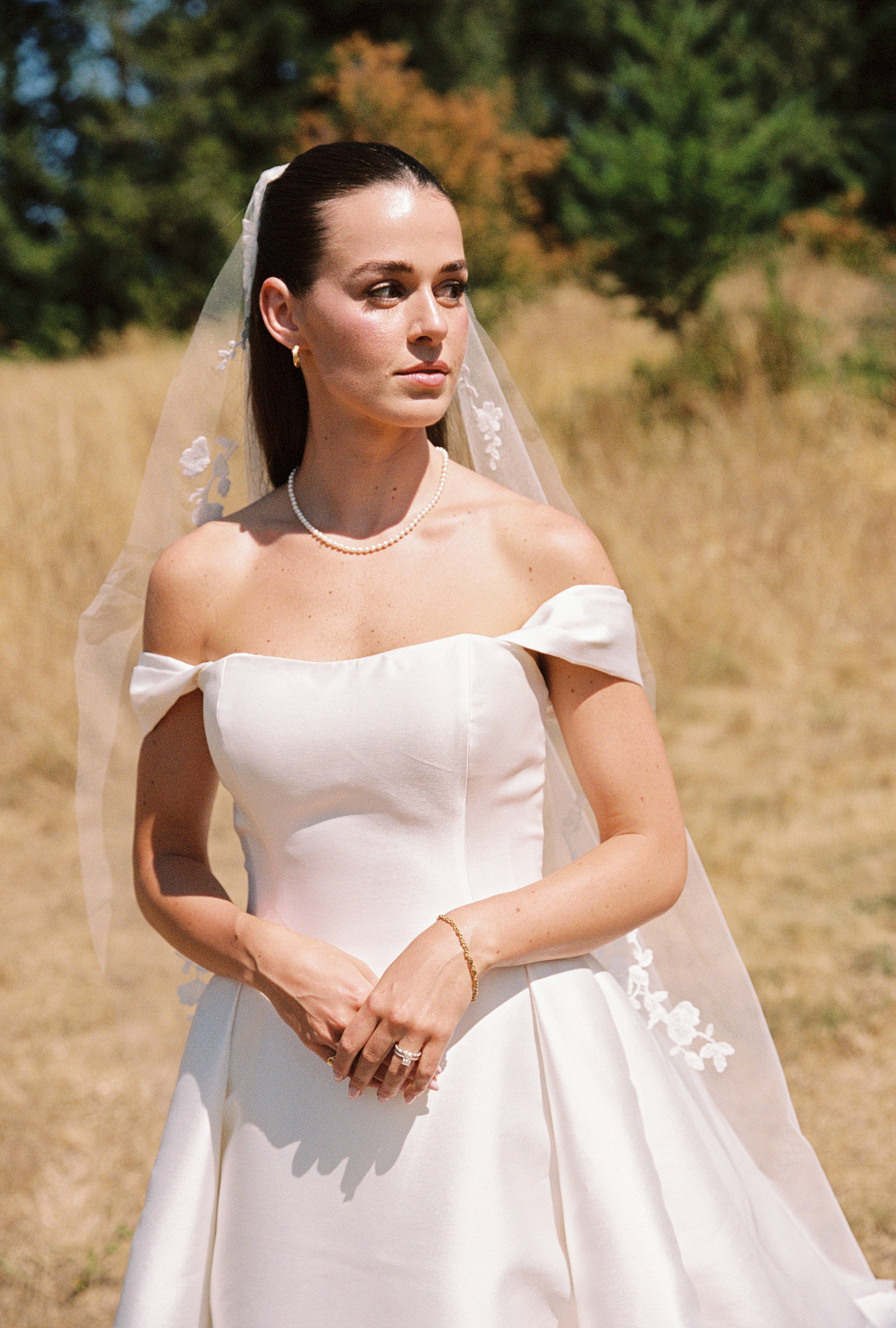 A woman in a white off-the-shoulder wedding dress standing outdoors in a grassy field with trees in the background.