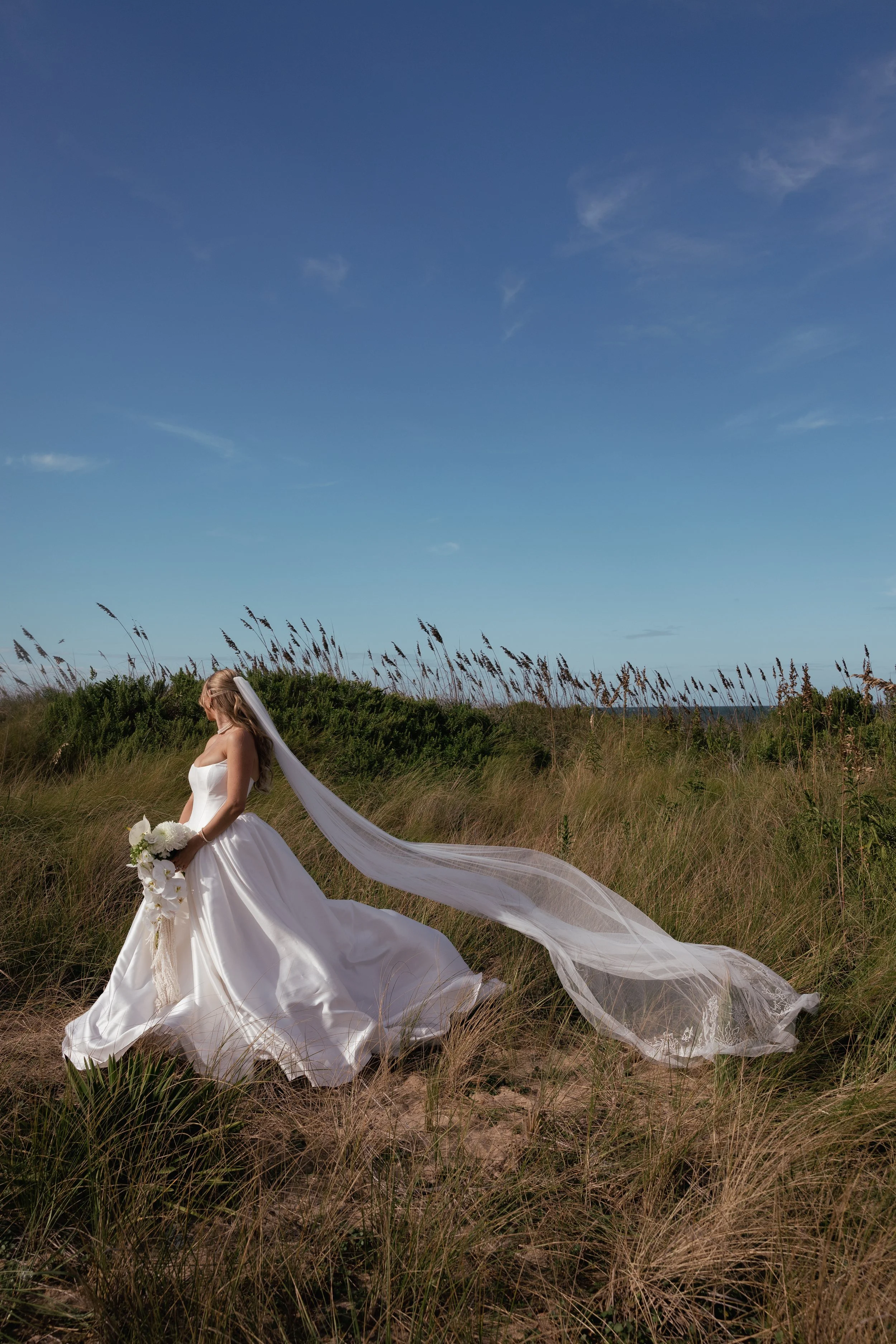 Bride in a white wedding gown holding a bouquet of flowers, standing in a grassy field under a clear blue sky with her veil flowing in the wind.