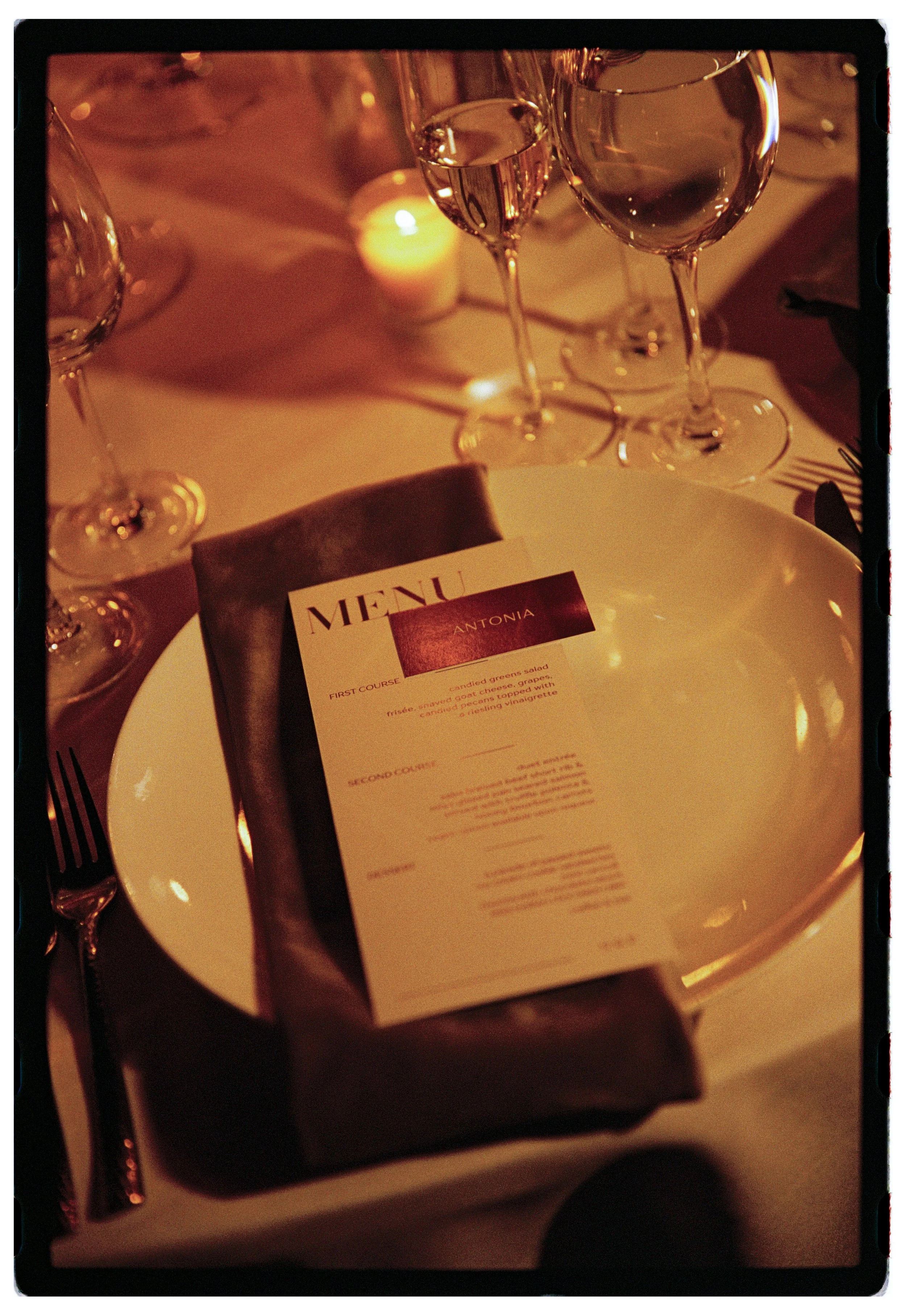 A table setting with a white plate holding a menu titled 'Antonia' and a dark napkin, surrounded by wine glasses and a lit candle in the background, creating an elegant dining atmosphere.