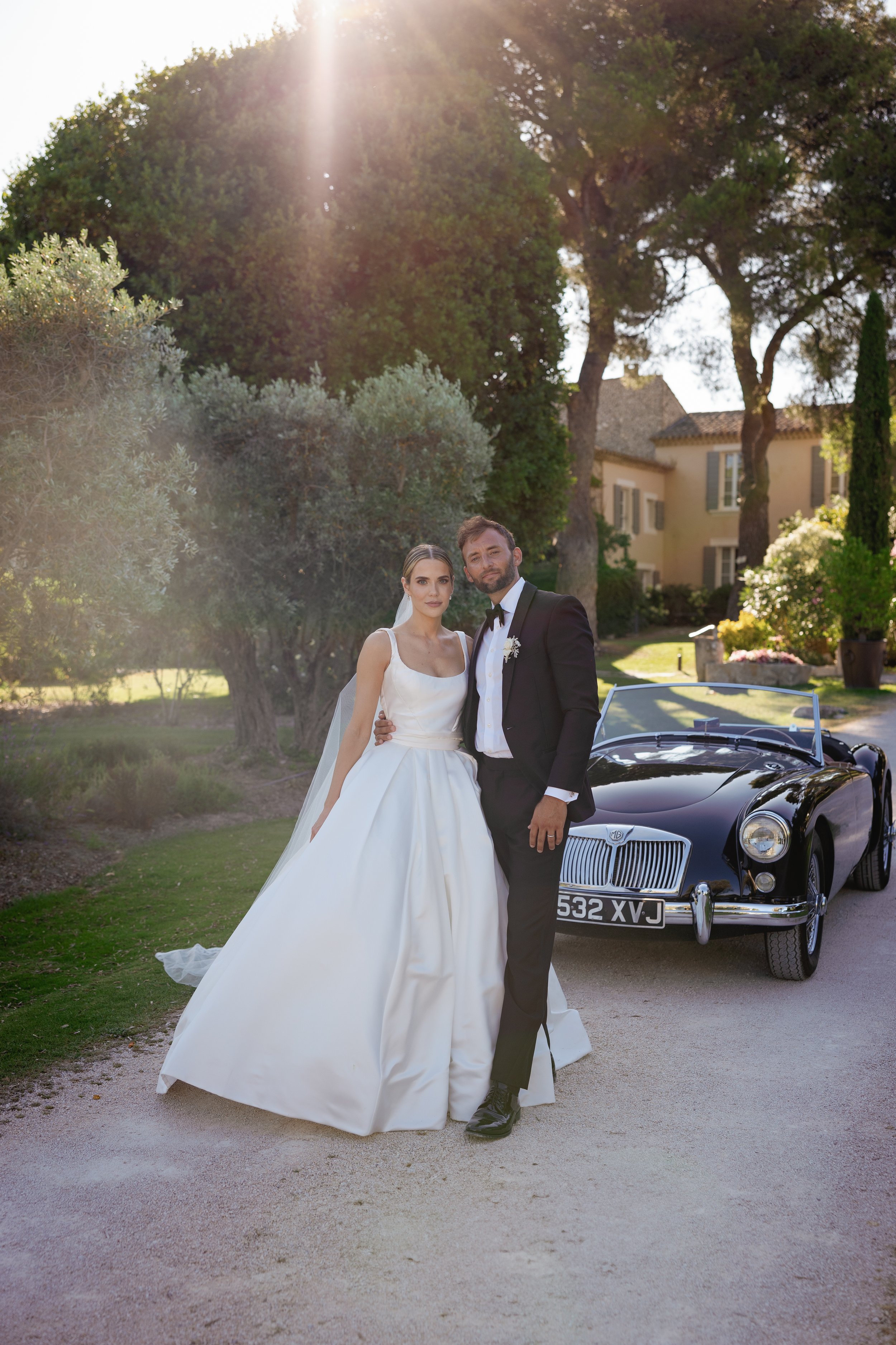 A bride and groom standing together in wedding attire outdoors next to a black vintage convertible car on a sunny day with trees and a house in the background.
