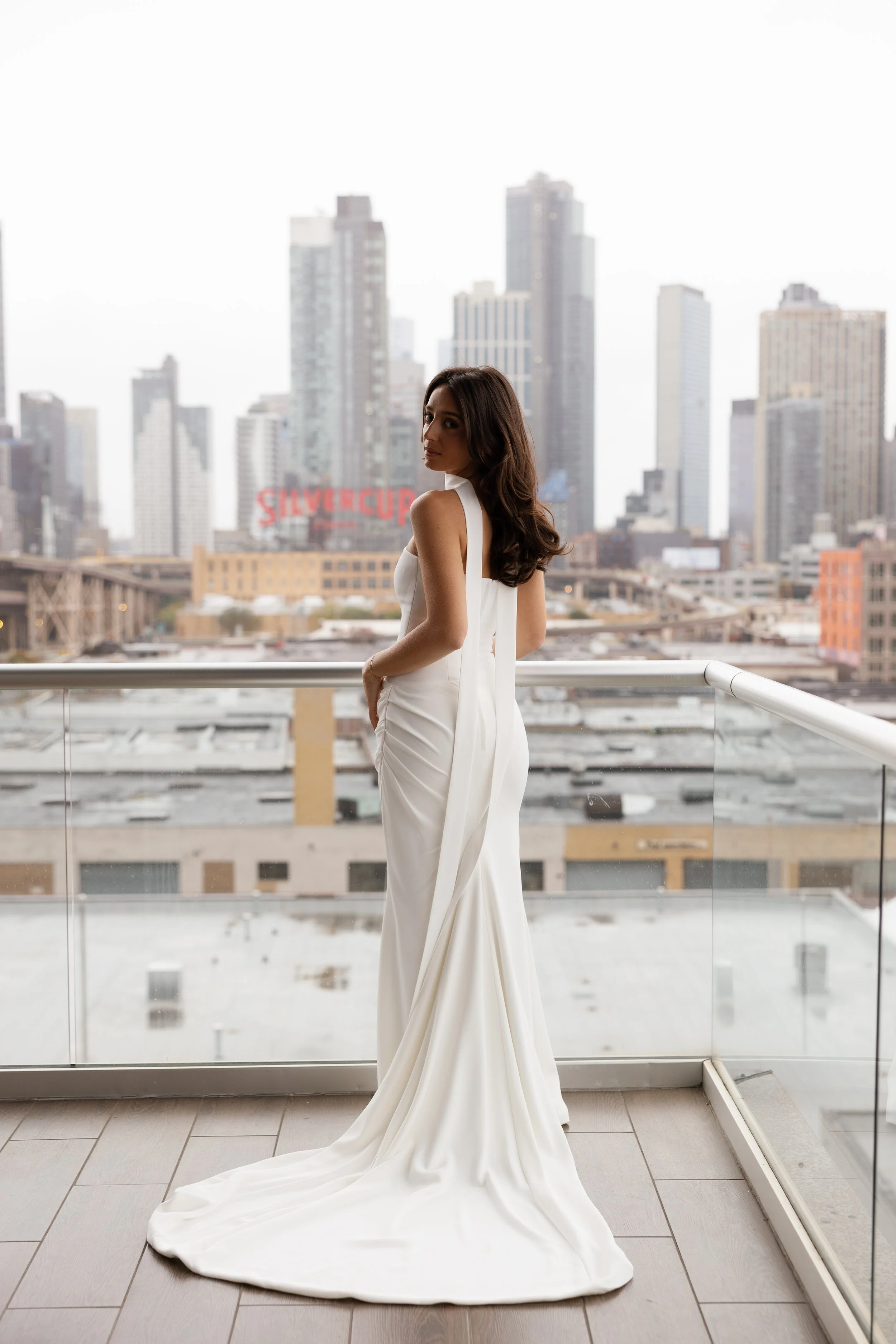 A woman in a white dress standing on a balcony with a city skyline in the background.