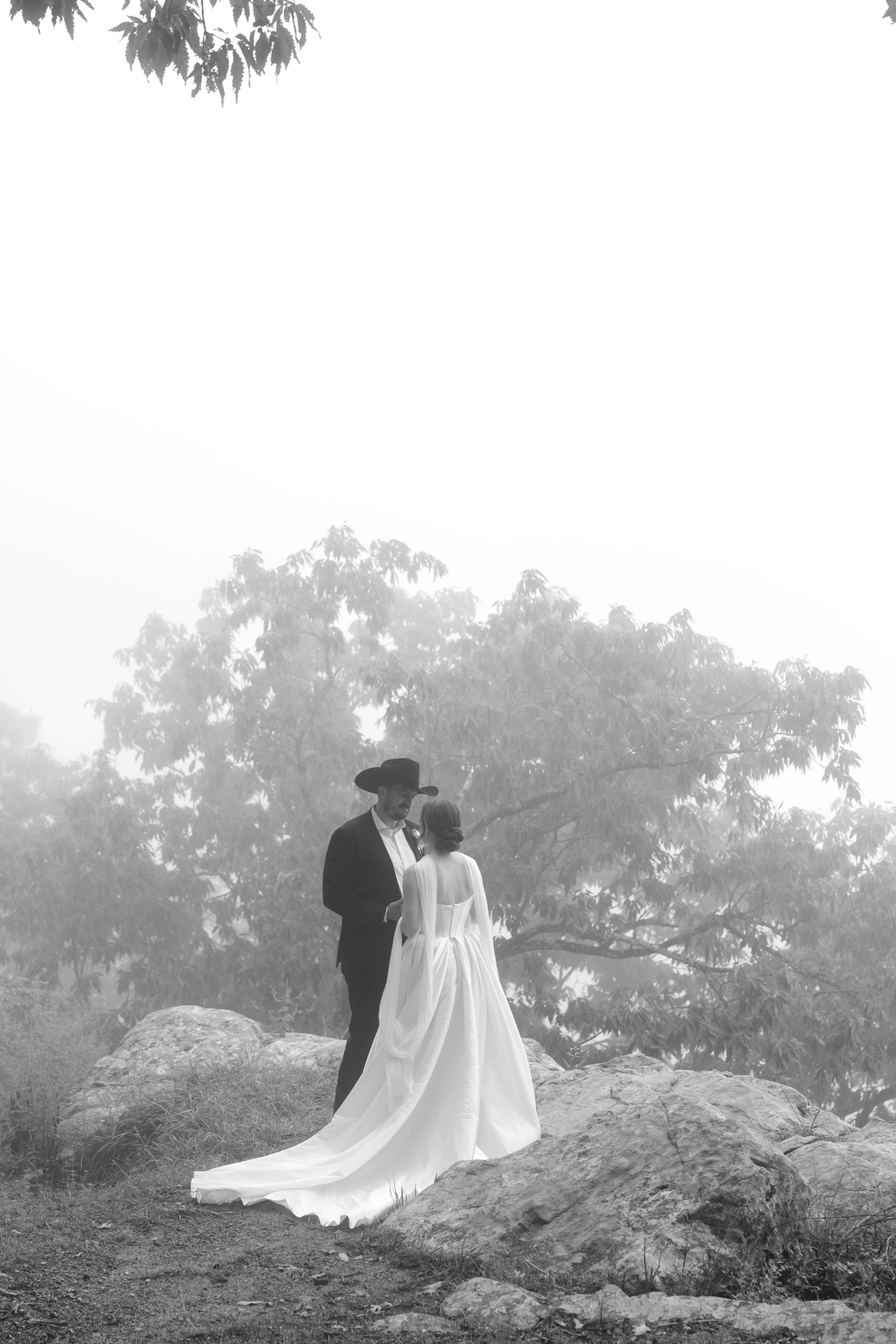 A black and white wedding photo of a man and woman dressed in wedding attire, standing on a rocky outdoor area with trees in the background, surrounded by mist or fog.