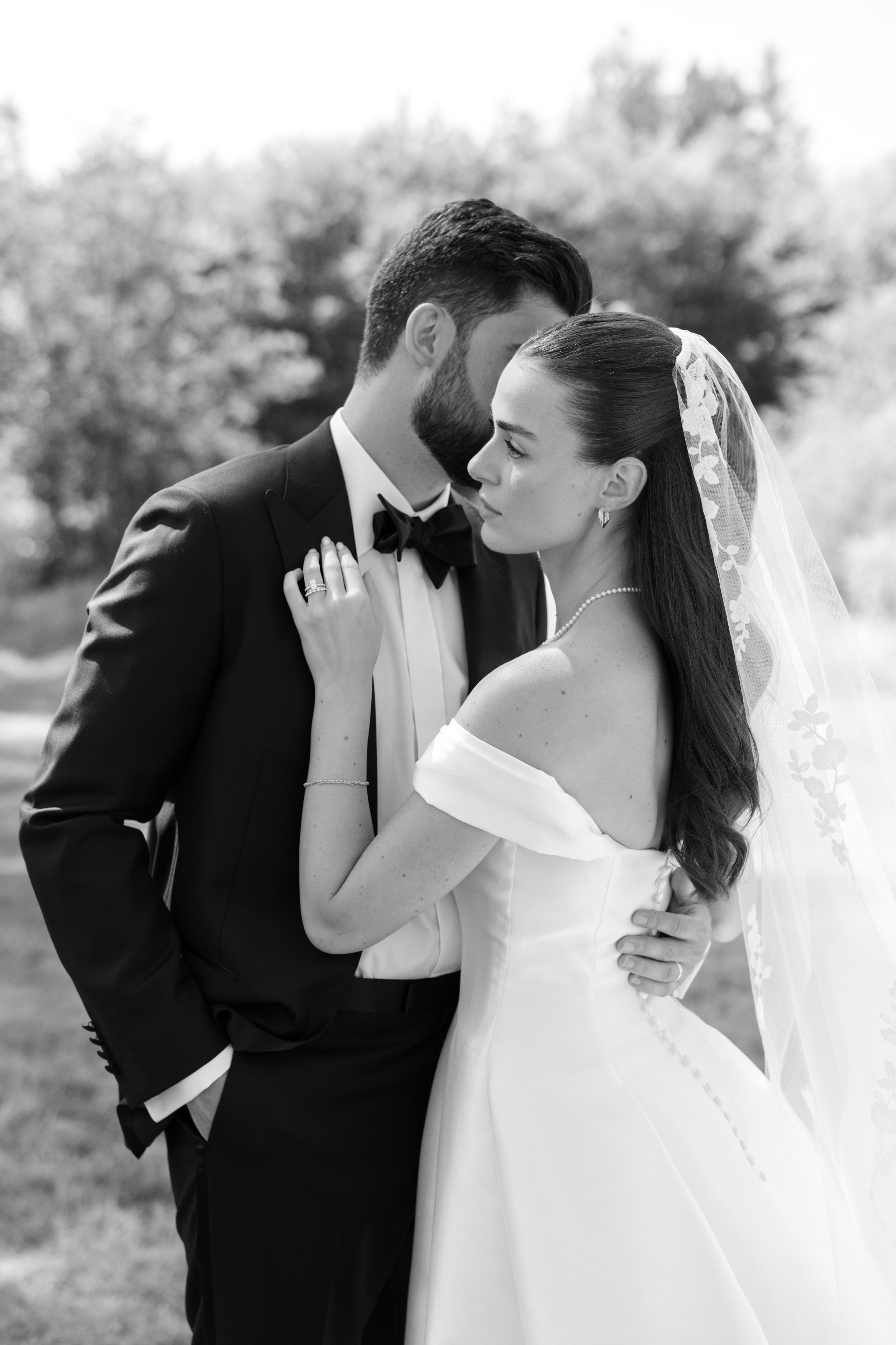 Black and white photo of a bride and groom embracing outdoors, with the bride touching the groom's face and the groom holding the bride's waist.
