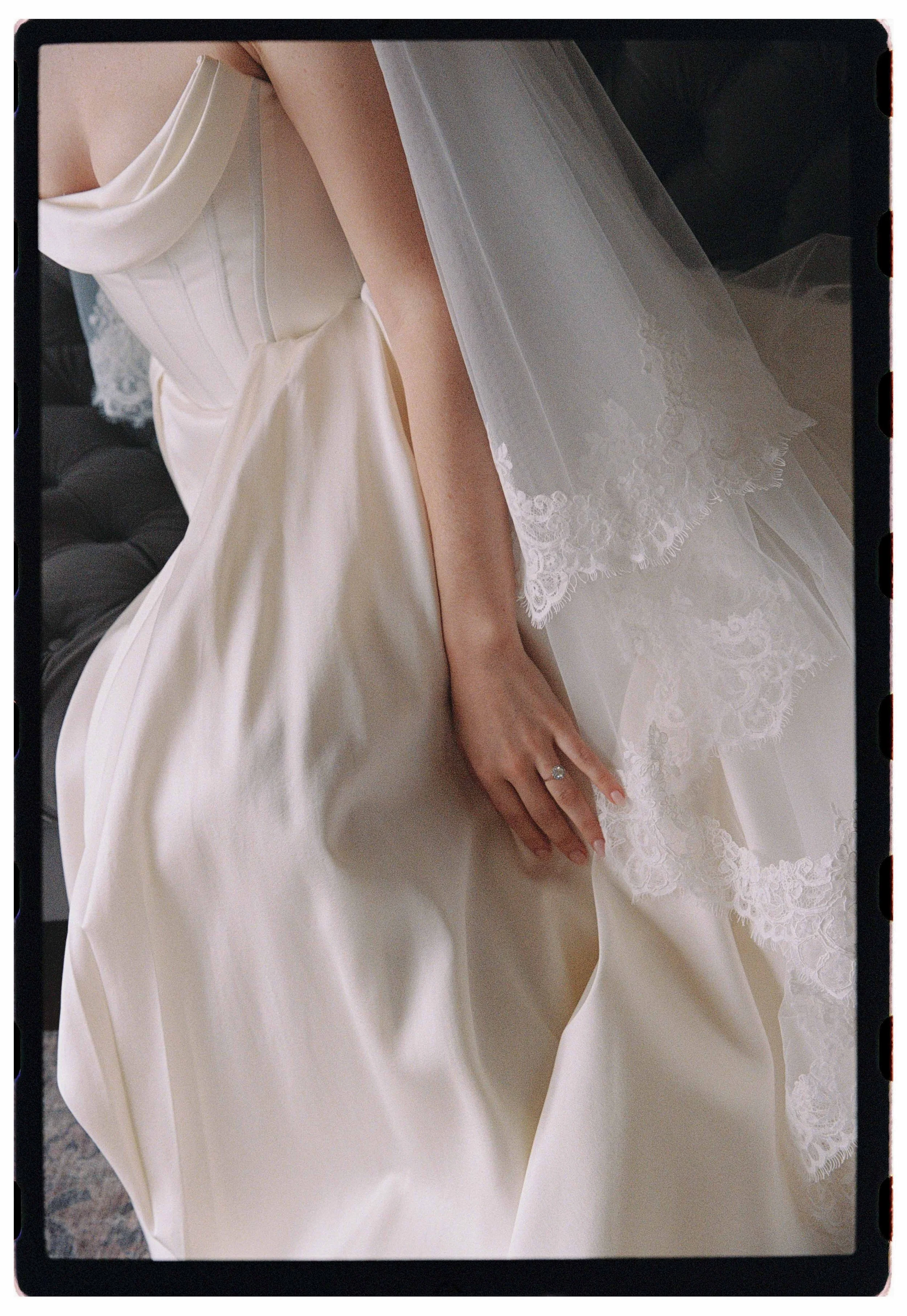 Close-up of a bride in a white satin wedding gown with lace details, sitting on a dark tufted sofa, showing her hand with a wedding ring.