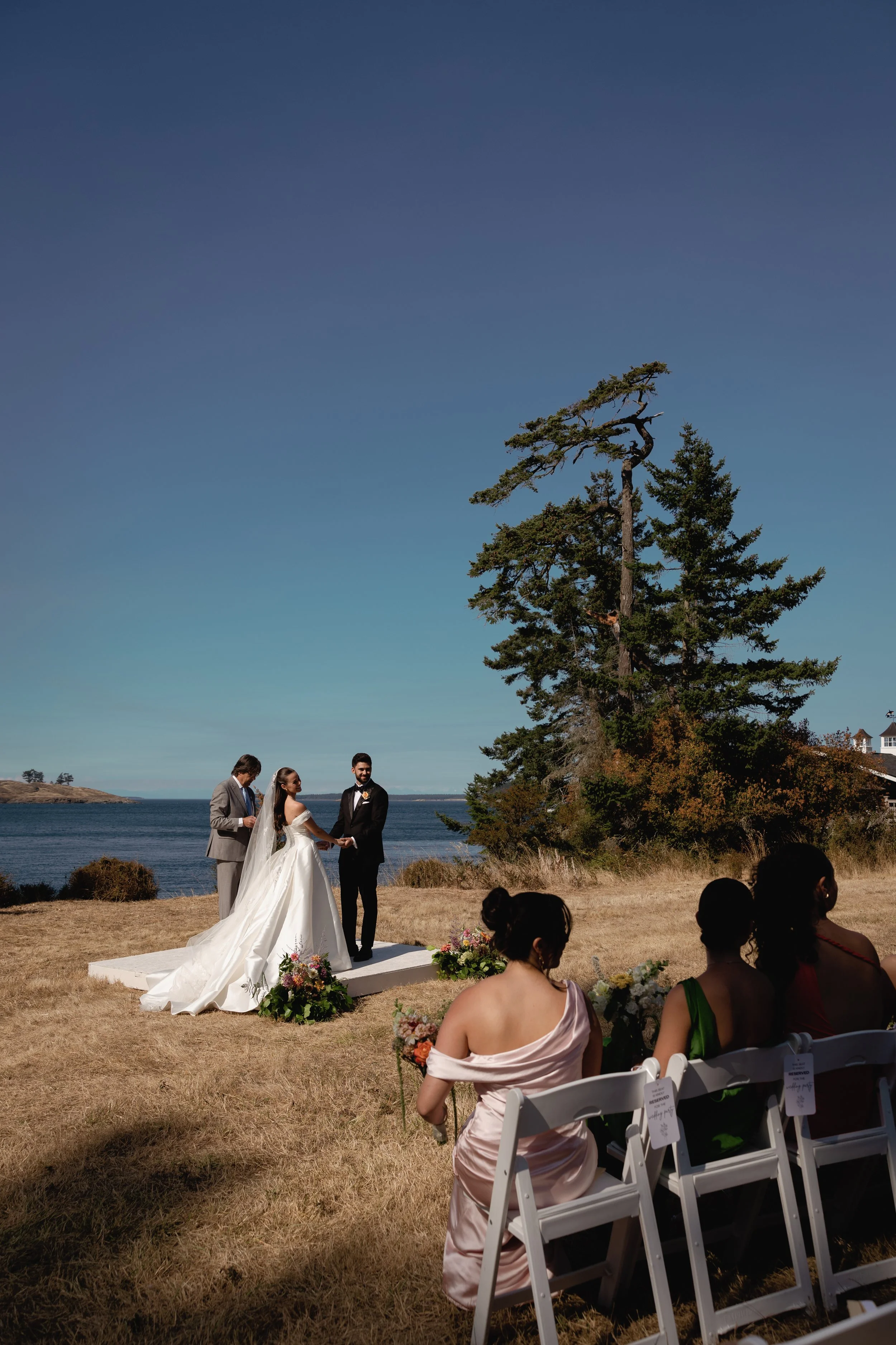 A couple getting married outdoors by the water with an officiant, surrounded by guests seated on white chairs, under a large tree on a sunny day.