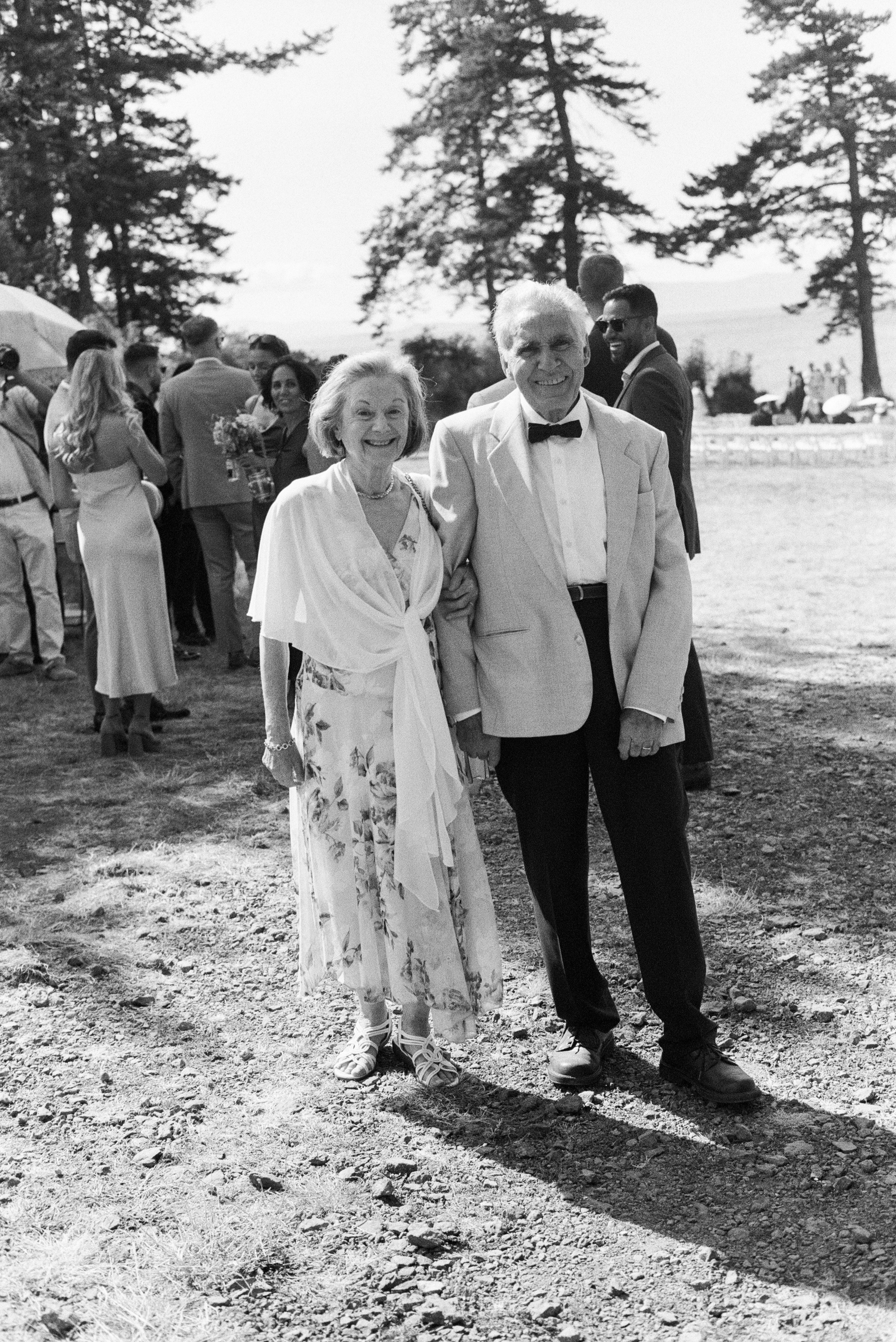 A black-and-white photo of an elderly couple smiling and holding hands at an outdoor event, with other people in the background. The woman wears a floral dress and a shawl, and the man wears a light-colored blazer and a bow tie.