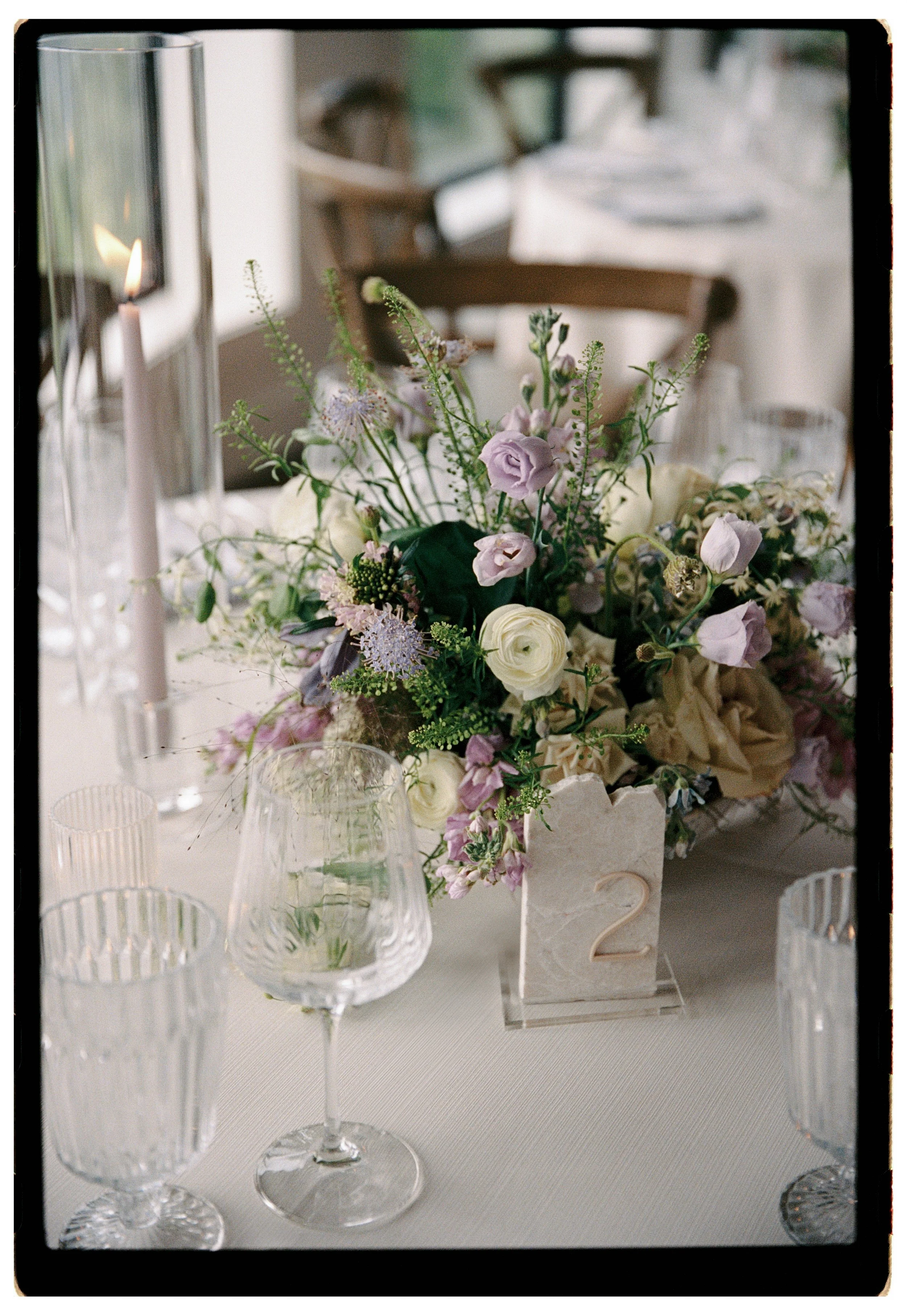 Elegant wedding table centerpiece with white and purple flowers, surrounded by empty glassware, with a candle in a tall glass holder on the side and a table number '2' in a decorative holder.