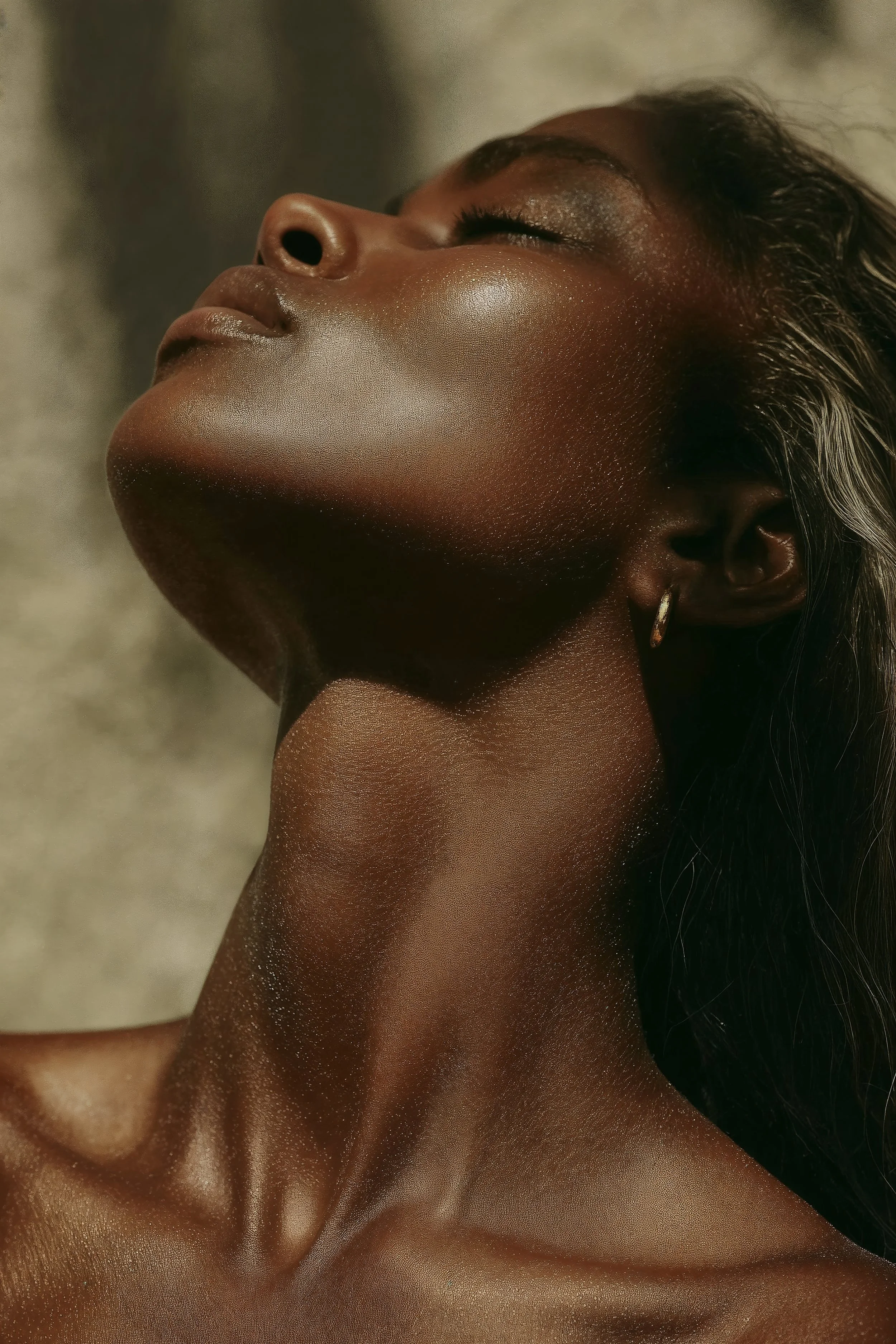 Close-up of a woman's face with closed eyes, focusing on her nose, lips, and skin texture. She has dark skin, makeup on her eyelids, and a small hoop earring visible in her ear.