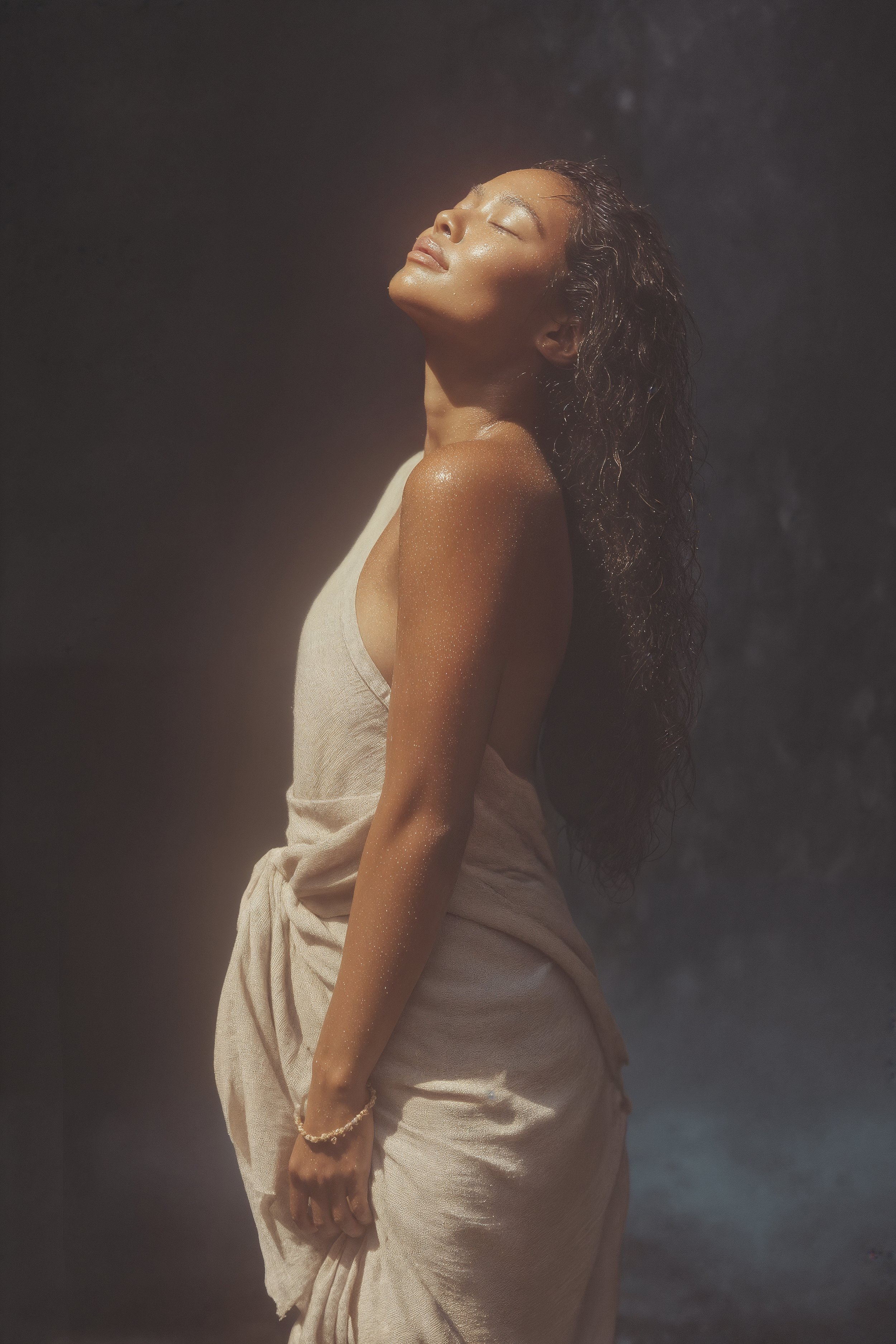 A brown-skinned woman with long wavy dark hair stands with her head titled up into the light, eyes closed,  a relaxed expression on her face, wearing a drapy sleeveless linen dress.