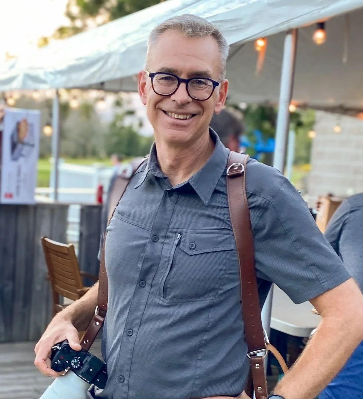 A smiling man in a gray button-up shirt and glasses holding a camera, standing outdoors at an event or gathering with a canopy and string lights in the background.
