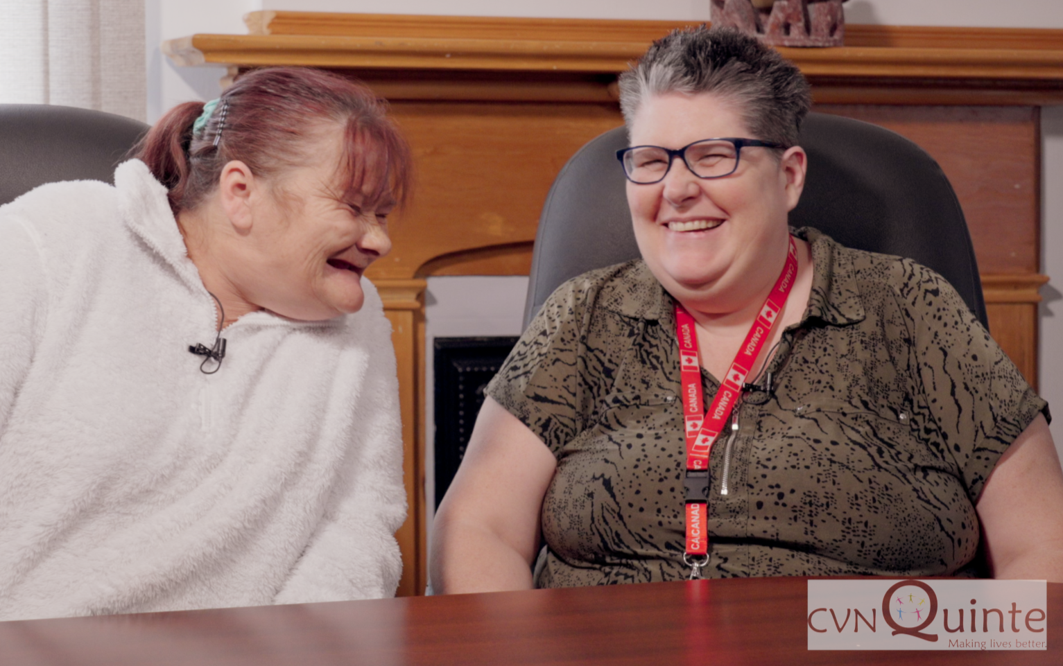 Two smiling women sitting at a table, one wearing a white fleece jacket and the other in a leopard print shirt with glasses and a red lanyard, in a room with wooden furniture.