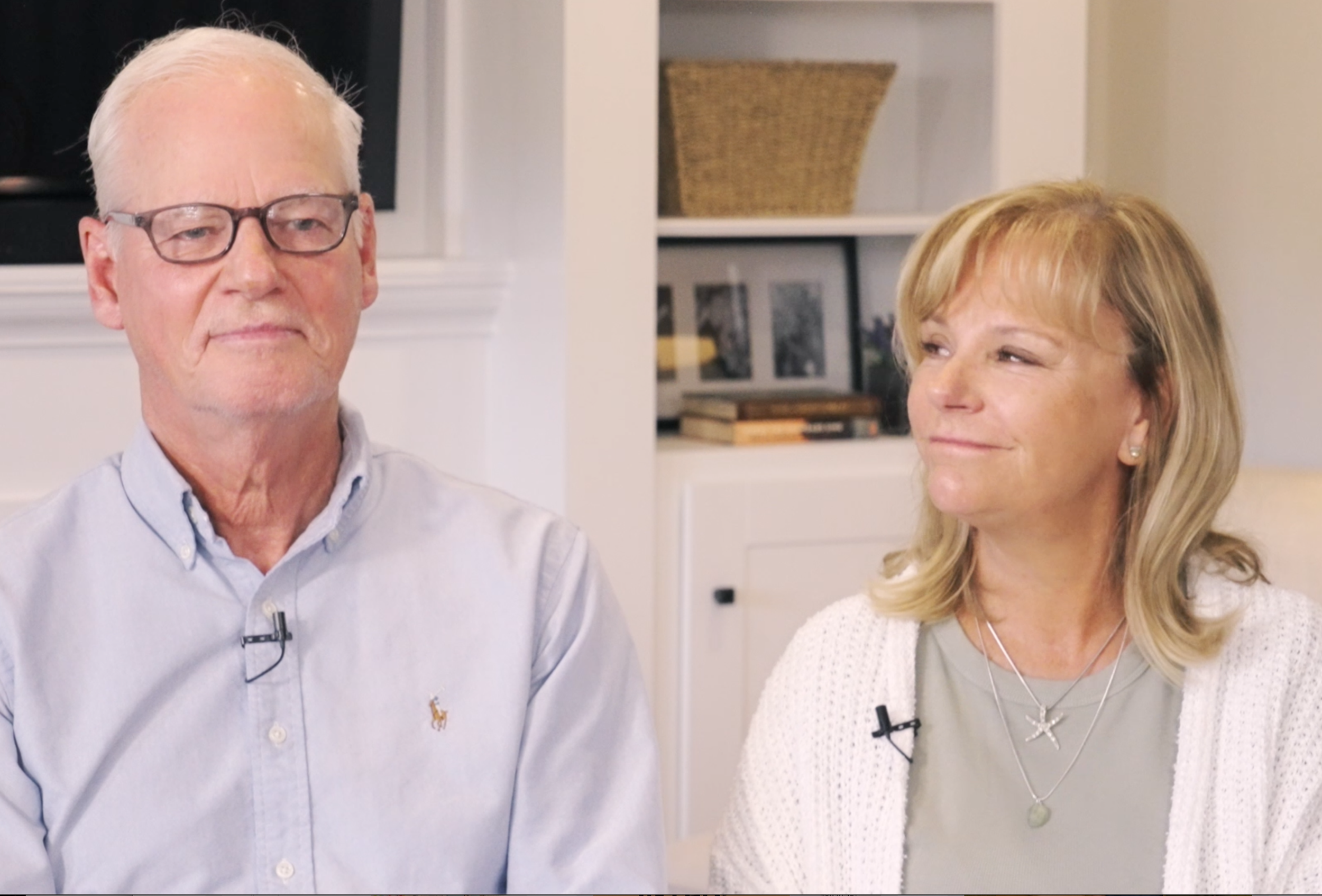A man with light hair and glasses, wearing a light blue button-down shirt with an embroidered logo, sitting next to a woman with blonde hair, wearing a gray top and a white cardigan with necklaces, in a cozy room with white shelves and decorative items in the background.