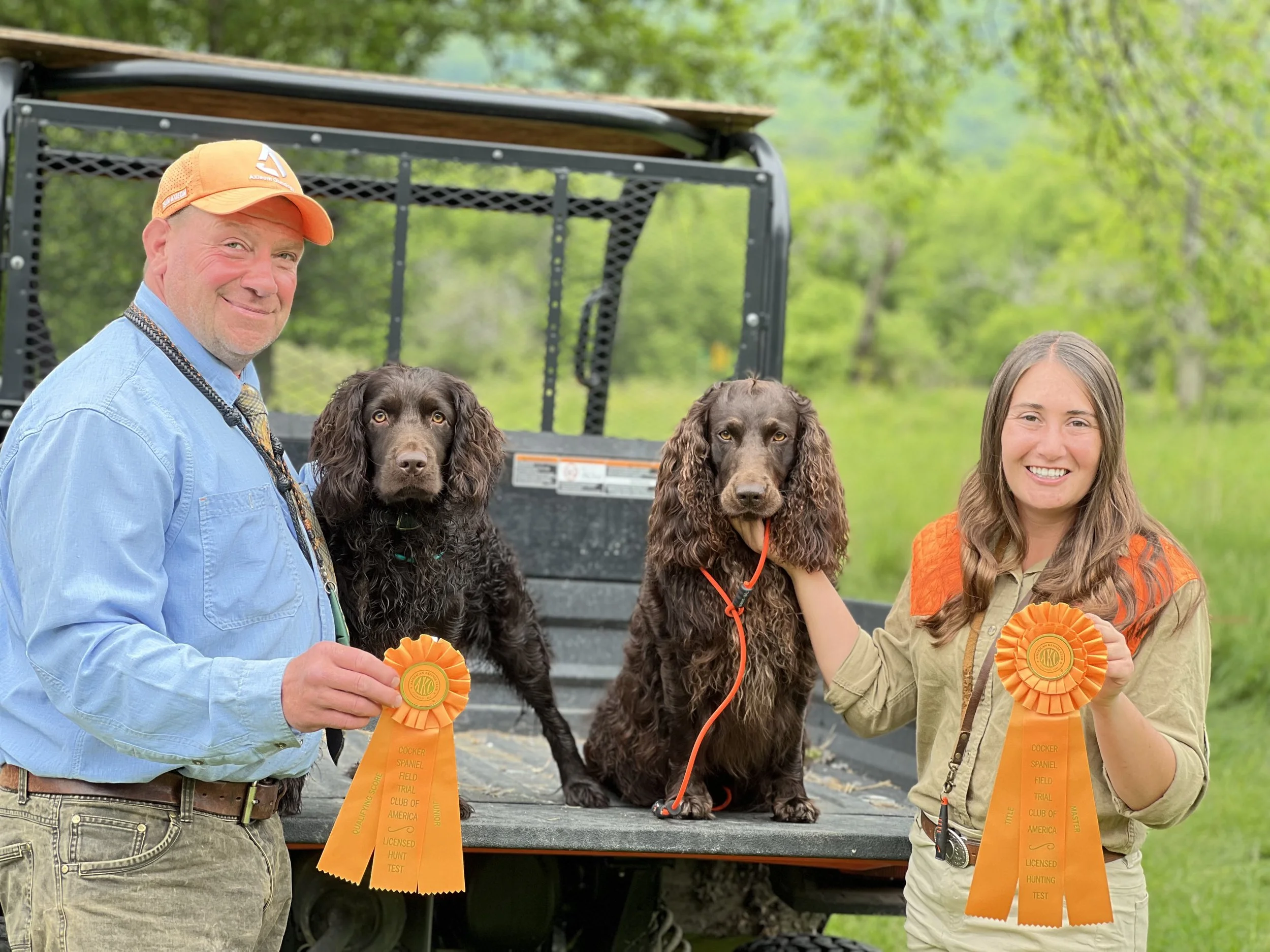 Two people, a man and a woman, are standing outdoors in front of a black utility trailer. They are holding orange ribbon awards. Between them are two spaniel dogs with brown fur and long ears. The man is wearing a light blue shirt, a patterned tie, a
