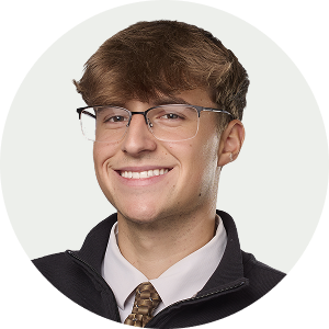 Photograph of a young man with light brown hair, glasses, and a friendly smile, wearing a blazer over a shirt and tie, against a light background.
