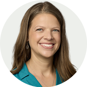 Headshot of a smiling woman with long brown hair, wearing a teal top and earrings, against a white background.
