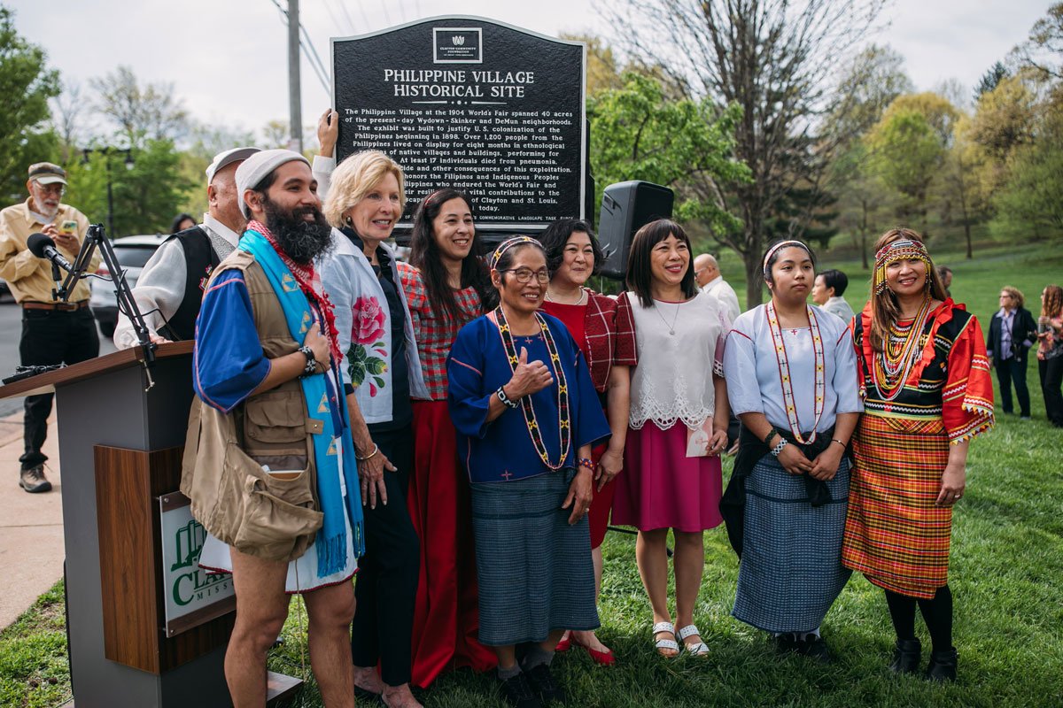 Group of diverse people in traditional and modern clothing at a historical site dedication ceremony, with a black informational plaque in the background.
