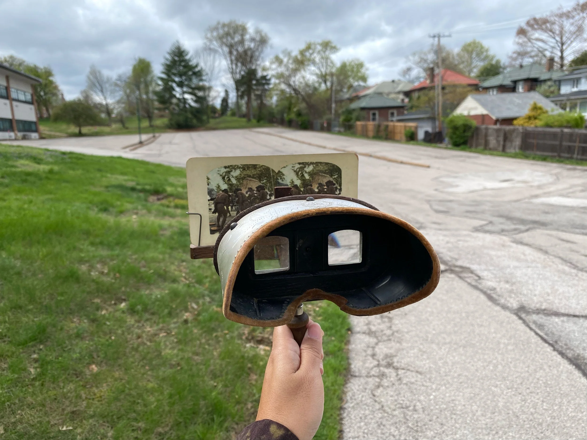 Person holding a vintage camera with a photo of soldiers in front of a parking lot and houses.