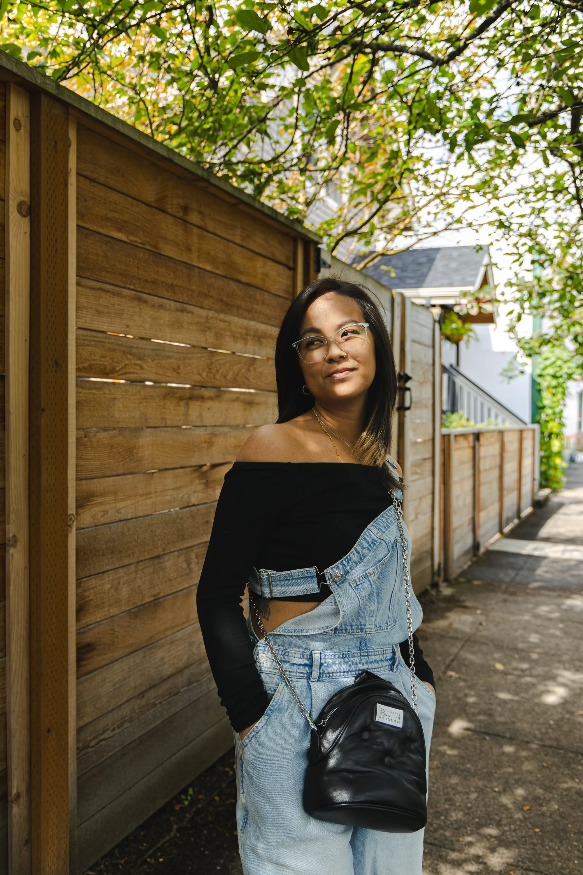 Person in black top and denim overalls standing by a wooden fence.