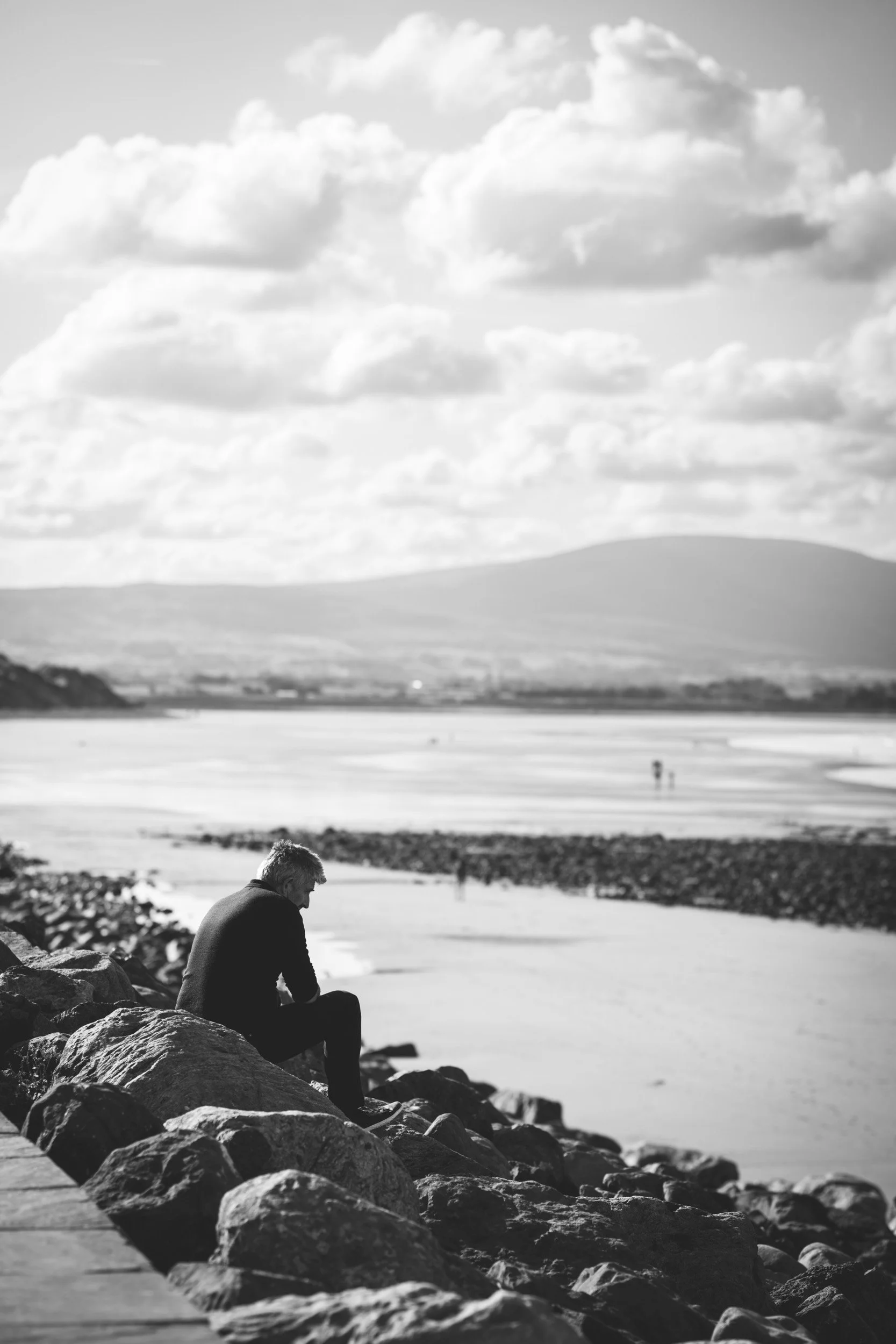 A black and white photo of a person sitting on rocks by a body of water, looking down, with hills and clouds in the background.