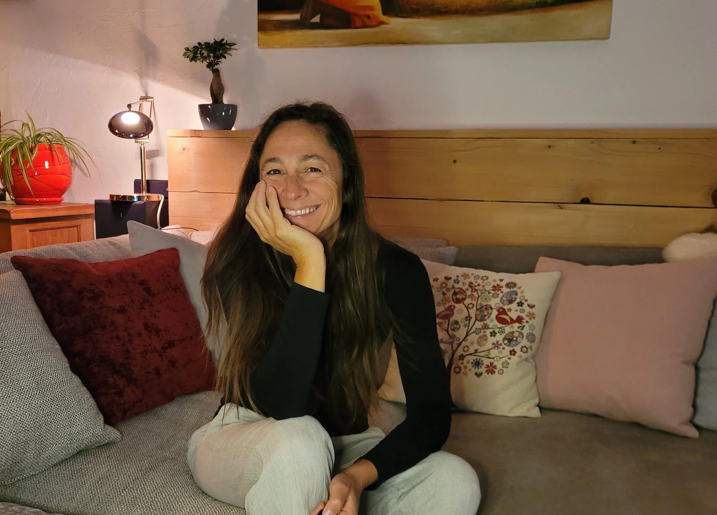 Transpersonal psychologist and spiritual teacher Kirsti smiles and rests her face on her hand, sitting on a beige sofa with various decorative pillows in a cosy living room.
