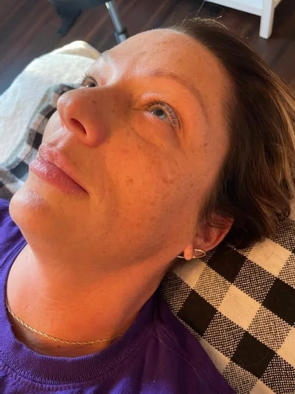A woman with short brown hair, wearing a purple shirt and gold earrings, lying on a plaid pillow. She has freckles and is looking upward, with a calm expression.