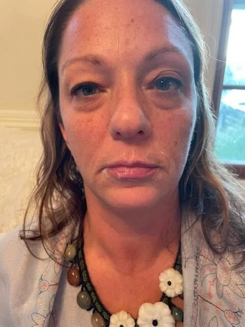Close-up of a woman with light skin and freckles, wearing a beaded necklace with white and brown flowers, indoors near a window.